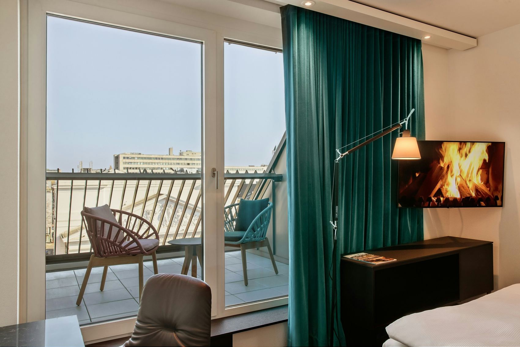 Hotel Motel One Aachen Modernes Hotelzimmer mit Balkon, zwei Stühlen und Tisch. Große Fenster bieten Tageslicht und Stadtblick. Gemütliche Atmosphäre durch grünen Vorhang und Fernseher mit Kaminfeuer.