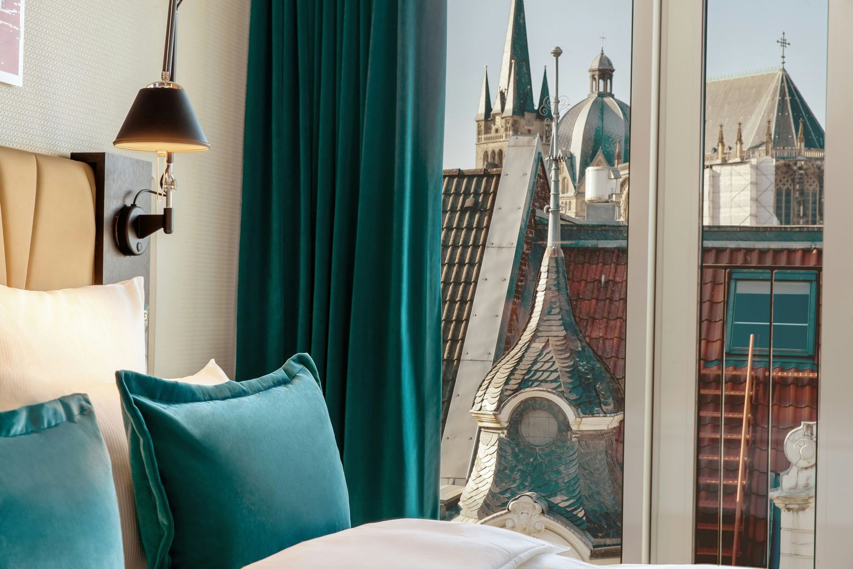 Hotel Motel One Aachen Blick aus einem Hotelzimmer mit bequemem Bett und türkisfarbenen Kissen auf historische Kirchturmspitzen und Dächer einer Altstadt bei klarem Himmel.