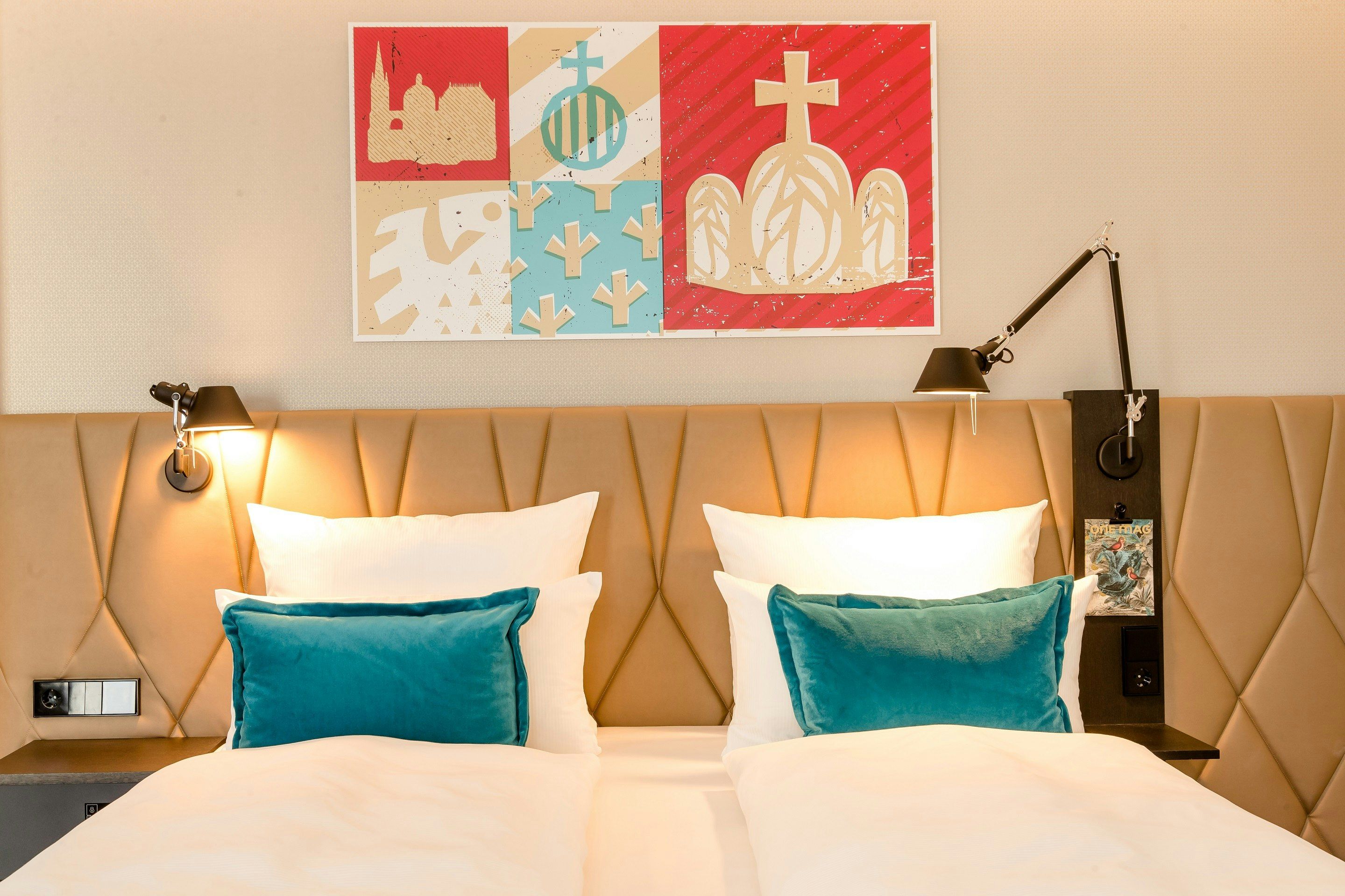 Hotel Motel One Aachen Modern eingerichtetes Doppelbett mit hellen Kissen und türkisfarbenen Zierkissen, sanfte Beleuchtung an der gepolsterten Wand sowie farbenfrohes, abstraktes Kunstwerk über dem Bett.