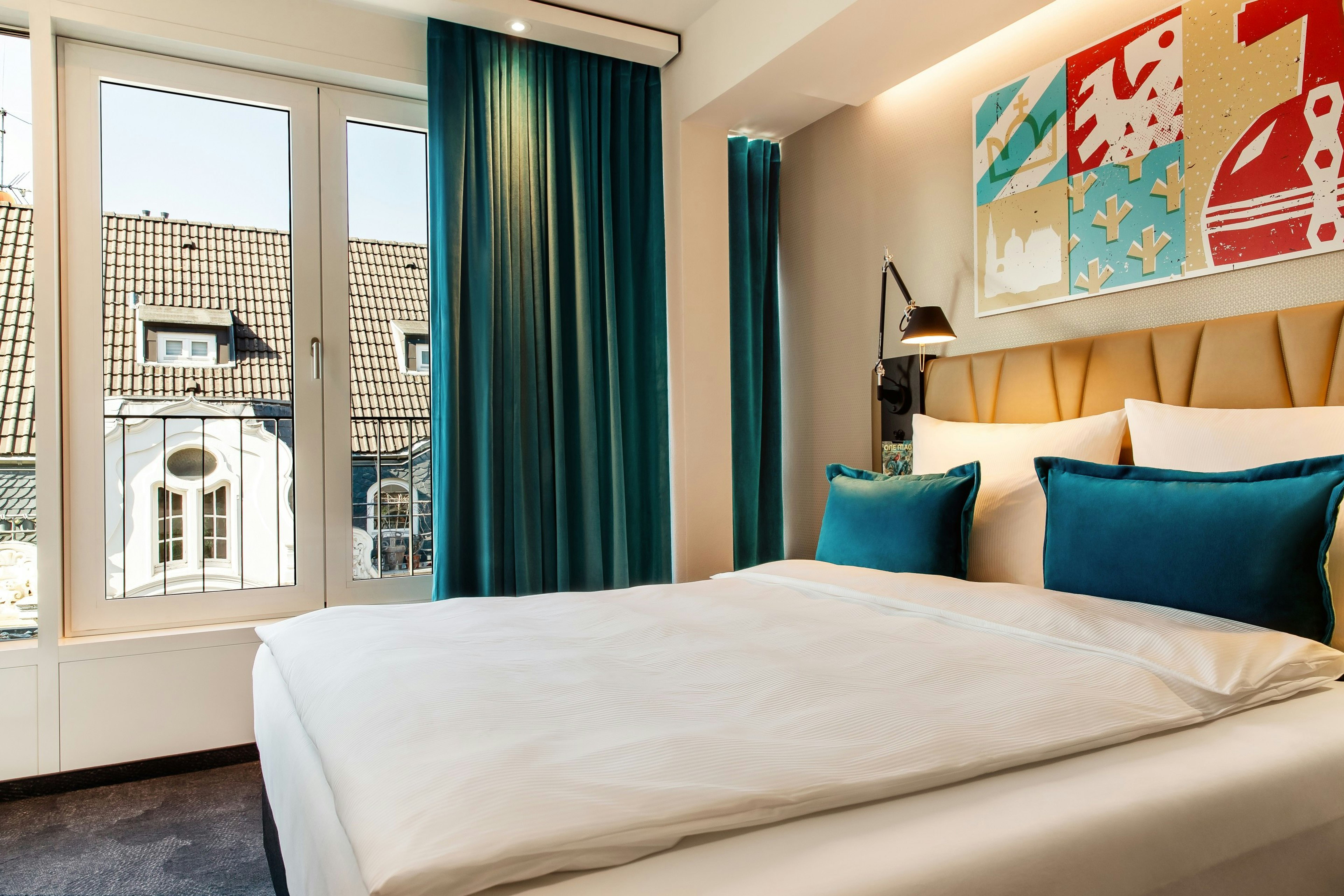 Hotel Motel One Aachen Helles Hotelzimmer mit großem Bett, türkisfarbenen Vorhängen und dekorativen Kissen. Großes Fenster bietet Blick auf traditionelle Dachlandschaft und sorgt für Tageslicht. Moderne, gemütliche Atmosphäre. Hotel Motel One Aachen Helles Hotelzimmer mit großem Bett, türkisfarbenen Vorhängen und dekorativen Kissen. Großes Fenster bietet Blick auf traditionelle Dachlandschaft und sorgt für Tageslicht. Moderne, gemütliche Atmosphäre.