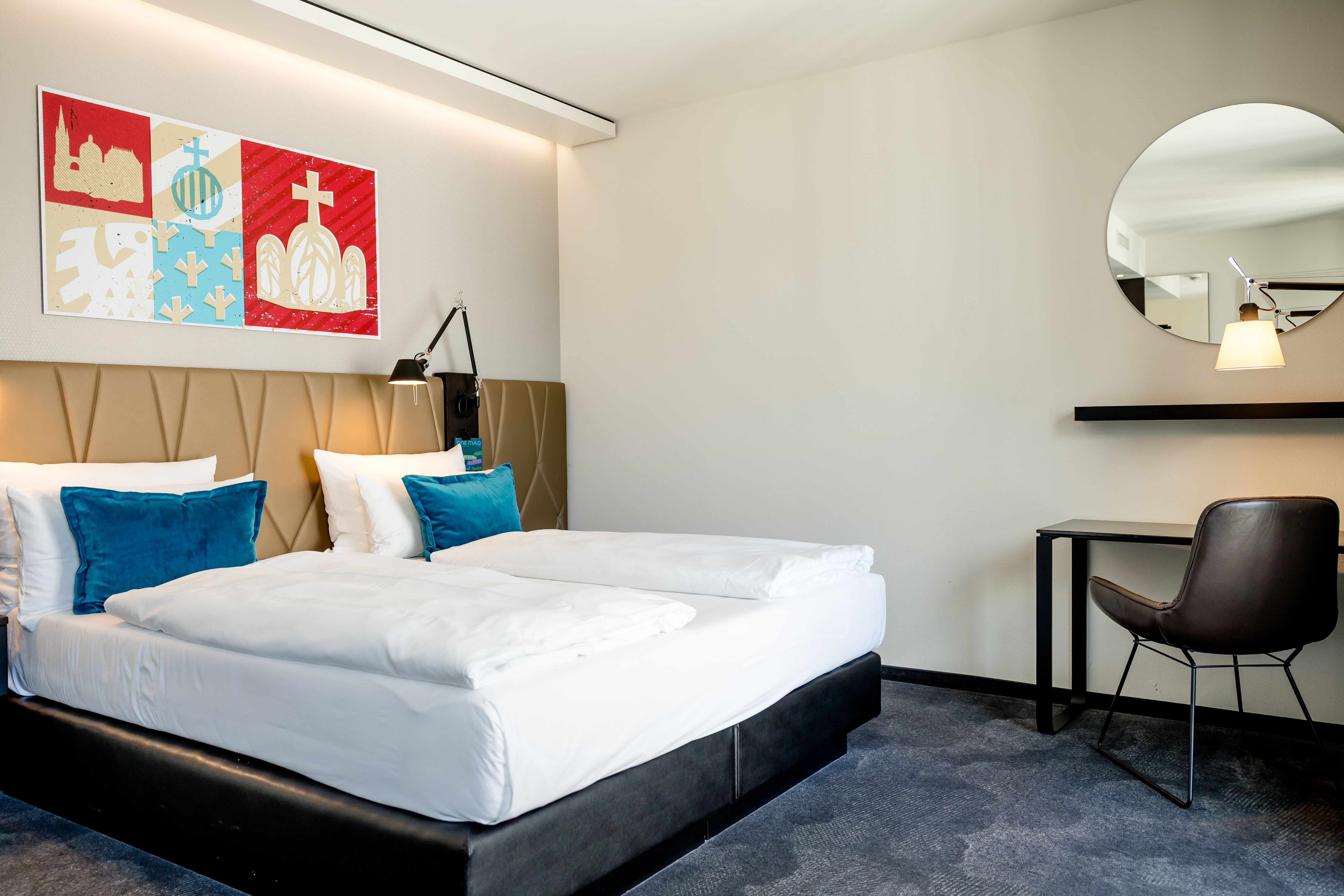 Modern eingerichtetes Hotelzimmer mit großem, gemütlichem Doppelbett, blauen Akzentkissen, hellem Ambiente und einer Arbeitsecke mit Schreibtisch, Stuhl und rundem Spiegel für Komfort und Funktionalität.