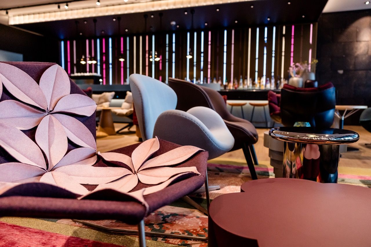 Moderne Lounge mit stilvollen Sesseln in unterschiedlichen Farben und weichen Textilien, dekorative Leuchtstäbe an der Wand, Atmosphäre wirkt einladend und komfortabel für entspannte Aufenthalte.