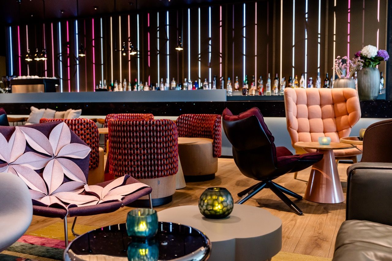 Moderne Hotelbar mit stilvollen Sesseln und gemütlicher Atmosphäre, gedämpftes Licht, unterschiedliche Sitzgruppen und dekorative Kerzen erzeugen einladendes Ambiente zum Entspannen und geselligen Beisammensein.