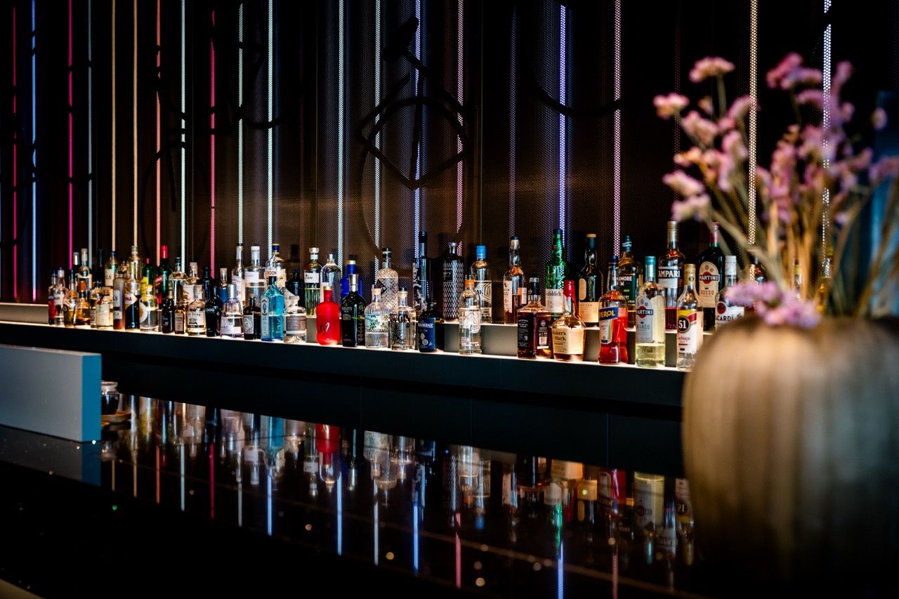 Moderne Hotelbar mit einer breiten Auswahl an Spirituosen auf schwarzer, glänzender Theke, beleuchtet durch dezente, farbige Lichtstreifen an der Wand, ergänzt durch eine Vase mit zarten Blüten im Vordergrund, die eine elegante und stilvolle Atmosphäre schafft.