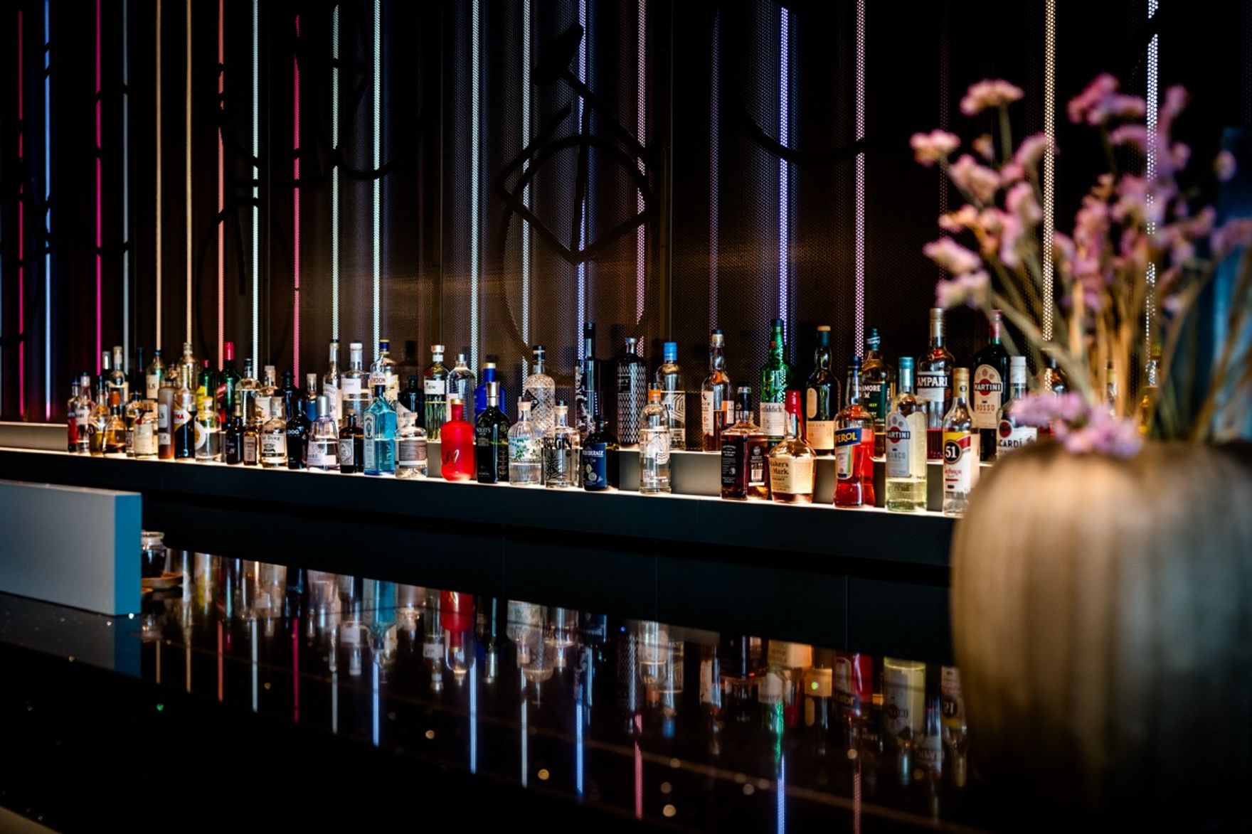 Moderne Hotelbar mit einer breiten Auswahl an Spirituosen auf schwarzer, glänzender Theke, beleuchtet durch dezente, farbige Lichtstreifen an der Wand, ergänzt durch eine Vase mit zarten Blüten im Vordergrund, die eine elegante und stilvolle Atmosphäre schafft.