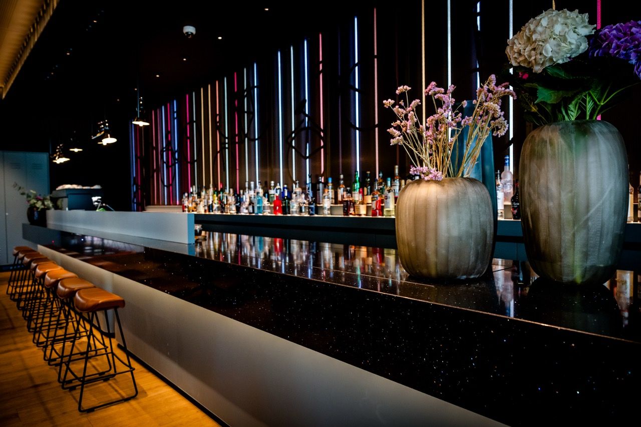 Moderne Hotelbar mit langer schwarzer Theke, stilvollen Blumenvasen und Hintergrundbeleuchtung in warmen Farben. Bequeme Barstühle laden zu entspannenden Momenten ein.