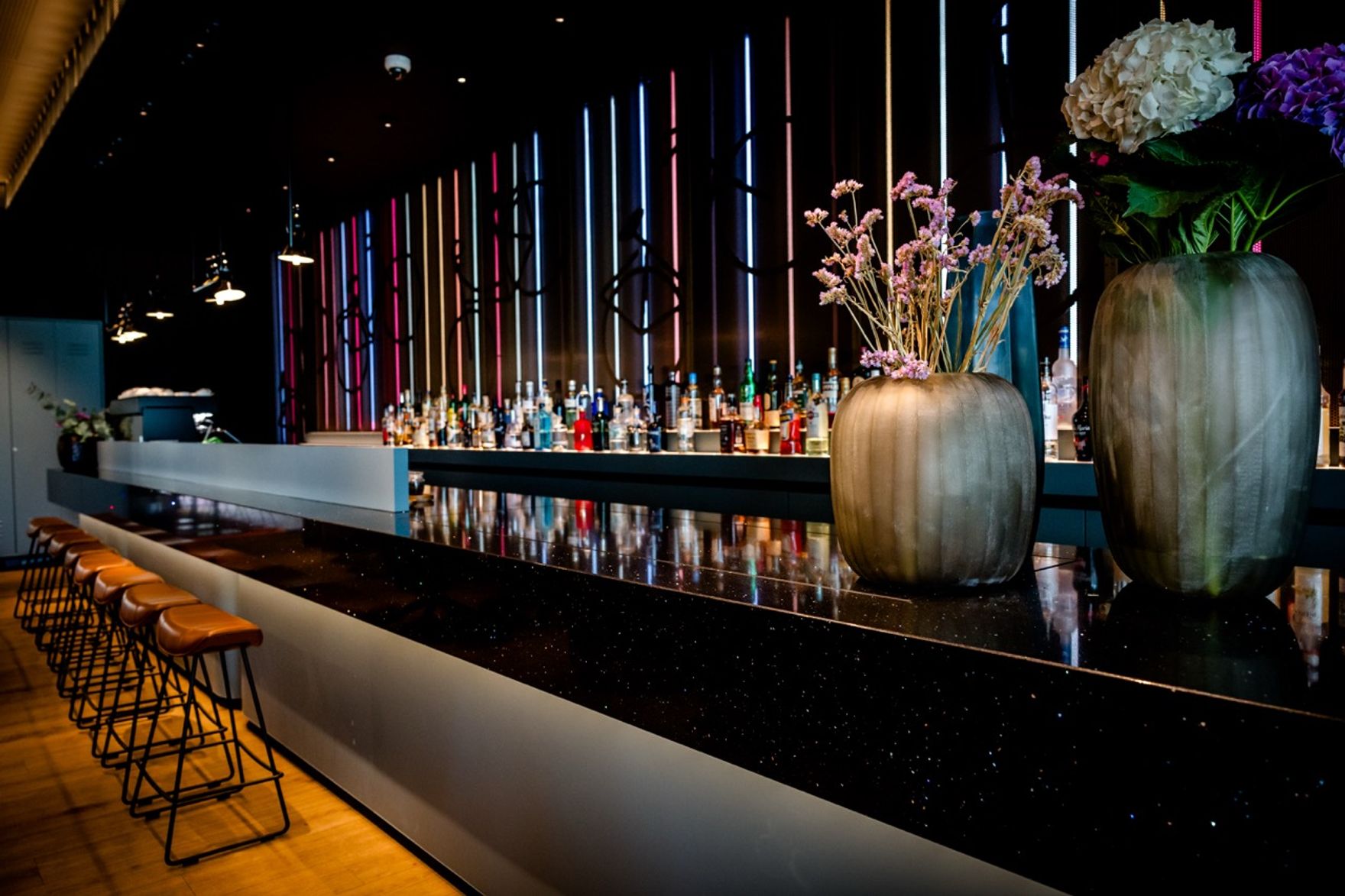 Moderne Hotelbar mit langer schwarzer Theke, stilvollen Blumenvasen und Hintergrundbeleuchtung in warmen Farben. Bequeme Barstühle laden zu entspannenden Momenten ein.