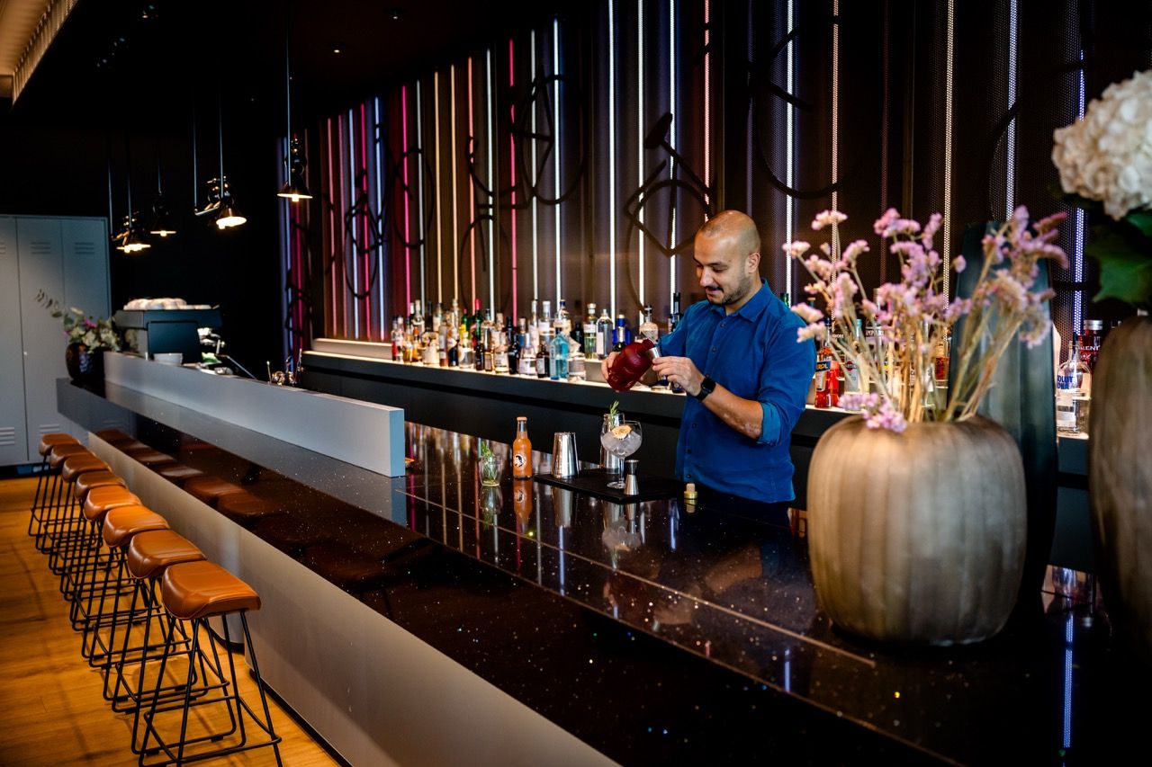 Barkeeper bereitet in einer stilvoll eingerichteten Hotelbar einen Cocktail zu. Die Bar ist modern gestaltet mit hohen Stühlen, stimmungsvoller Beleuchtung und dekorativen Blumenarrangements.