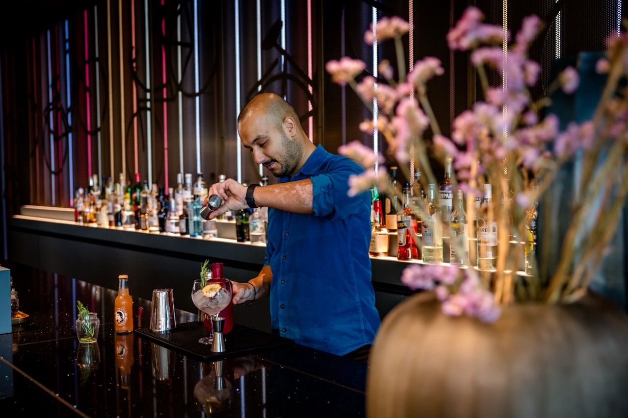 Ein Barkeeper bereitet hinter einer modernen Bar einen Cocktail zu, umgeben von einer vielfältigen Auswahl an Spirituosen. Im Vordergrund sind dekorative, rosa Blüten unscharf zu sehen.