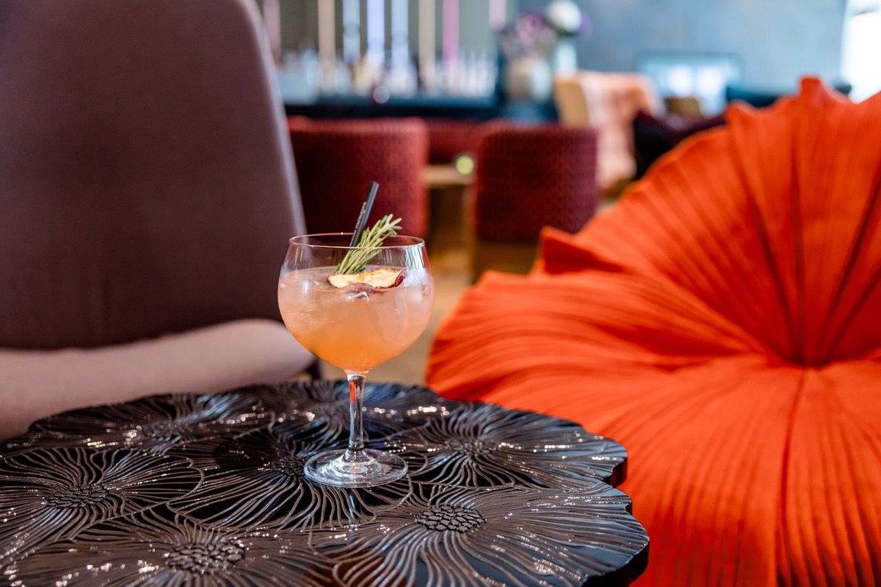 Cocktailglas mit erfrischendem Getränk und Kräuterzweig steht auf einem kunstvoll gestalteten schwarzen Tisch, im Hintergrund gemütliche, farbige Sitzmöbel in warmem Ambiente einer Bar oder Lounge.