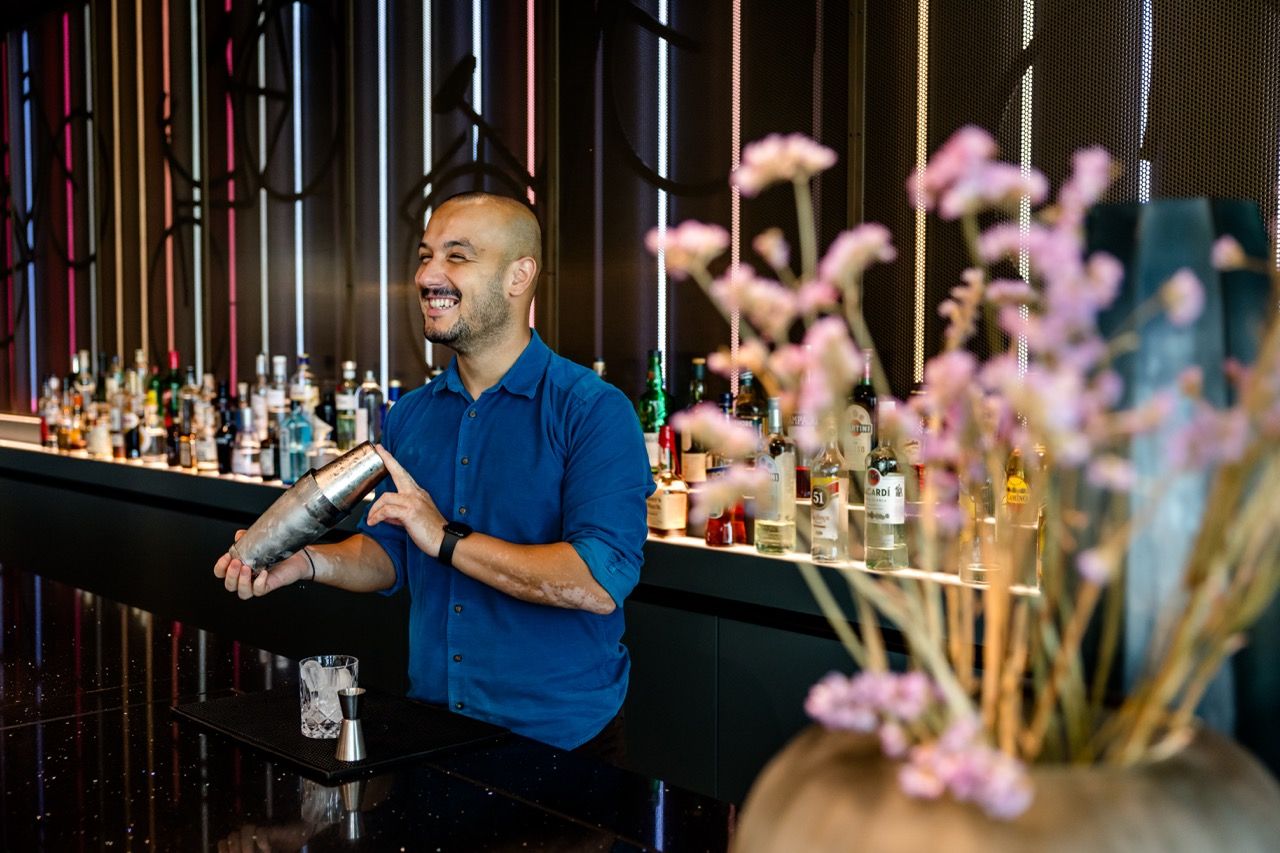 Ein Barkeeper schüttelt fröhlich einen Cocktail-Shaker hinter einer gut ausgestatteten Bar mit vielen Spirituosenflaschen, umgeben von moderner Einrichtung und sanfter Beleuchtung. Im Vordergrund unscharfe Blumen sorgen für eine einladende Atmosphäre.