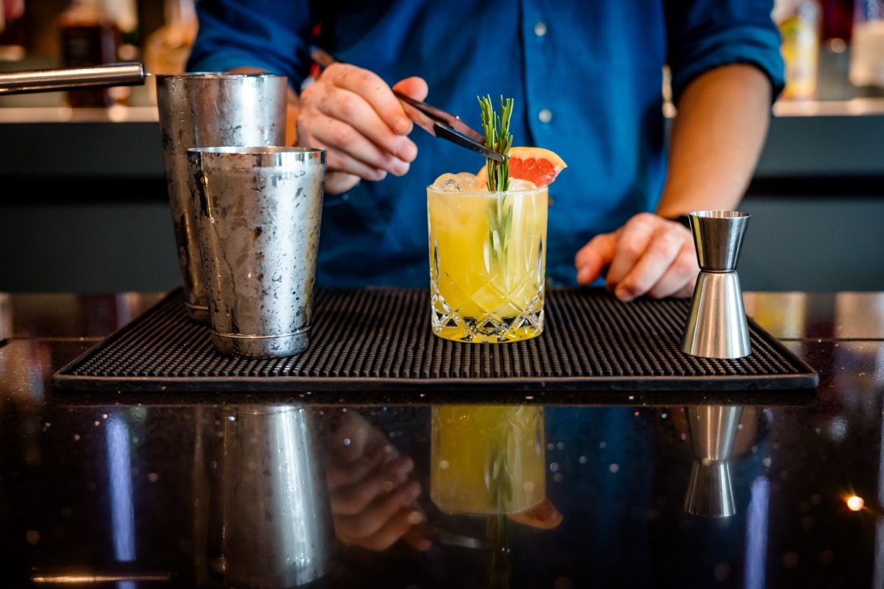 Barkeeper dekoriert einen frisch zubereiteten, gelben Cocktail mit einem Zweig Rosmarin und einer Grapefruitscheibe an einer stilvollen Hotelbar.
