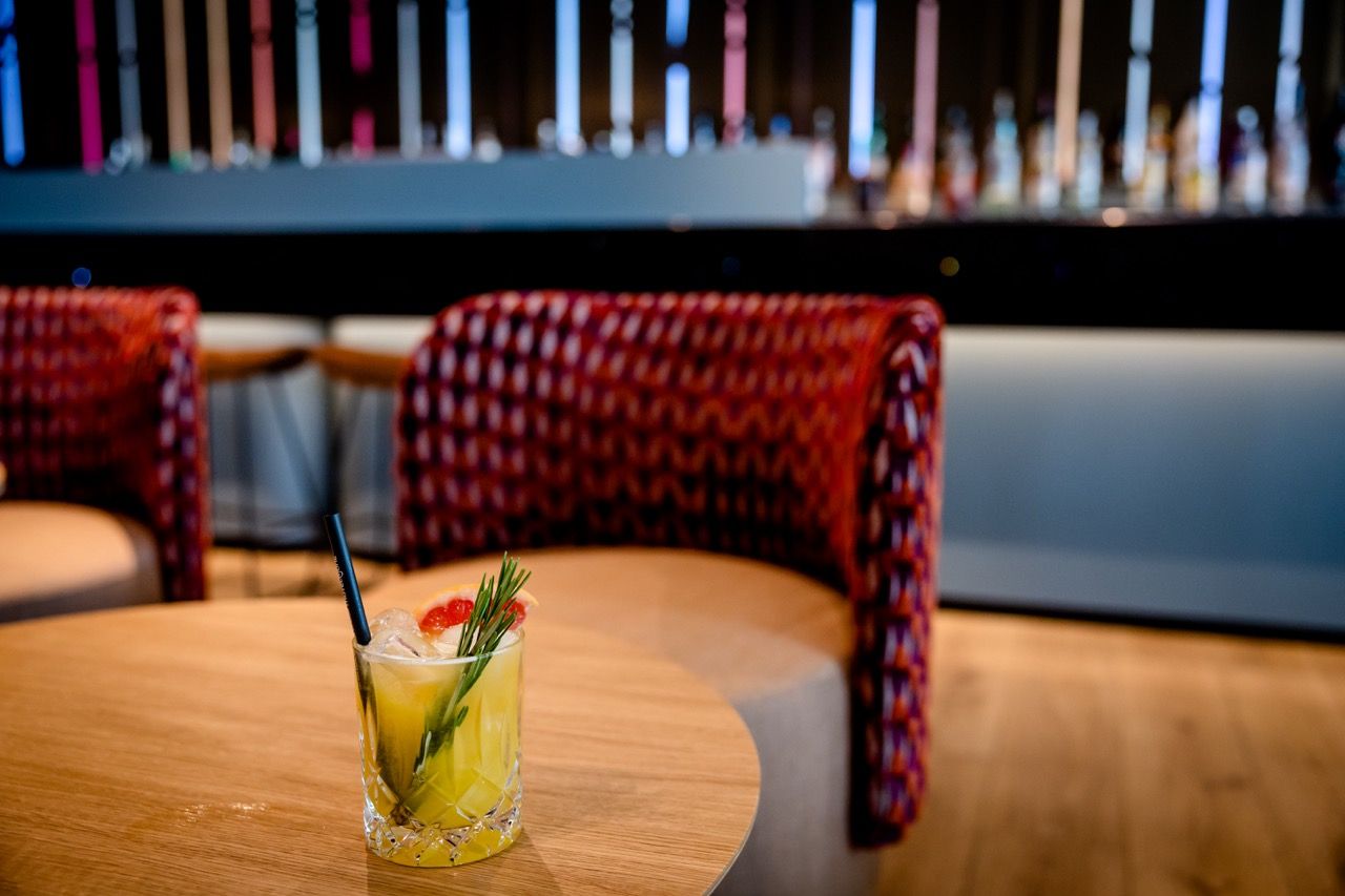 Ein erfrischender Cocktail mit Garnitur steht auf einem Holztisch in einer stilvollen Hotelbar mit gemütlichen, roten Sesseln im Hintergrund.