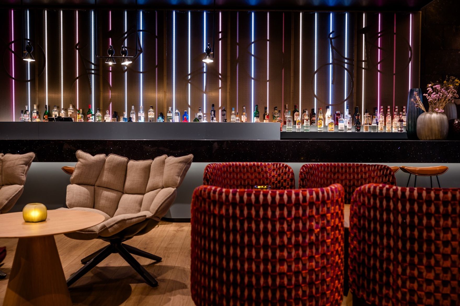 Moderner Hotelbarbereich mit gemütlichen Sesseln, warmem Licht und stilvoll beleuchteter Rückwand, an der verschiedene Spirituosen präsentiert werden. Einladende Atmosphäre zum Entspannen und Genießen.