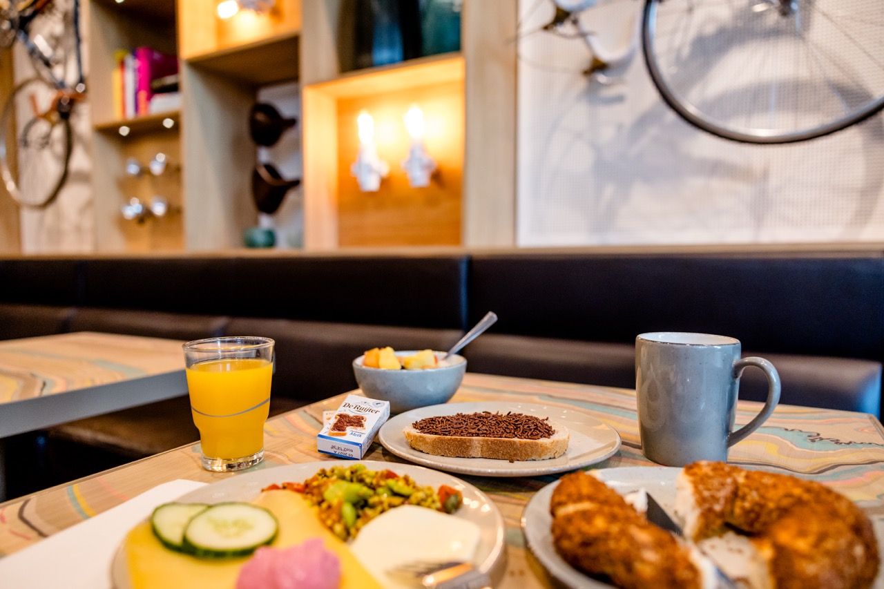 Frühstückstisch mit frisch gepresstem Orangensaft, Müsli, Kaffee, Brot mit Schokostreuseln, Aufschnitt und Gebäck in gemütlichem Café-Ambiente.