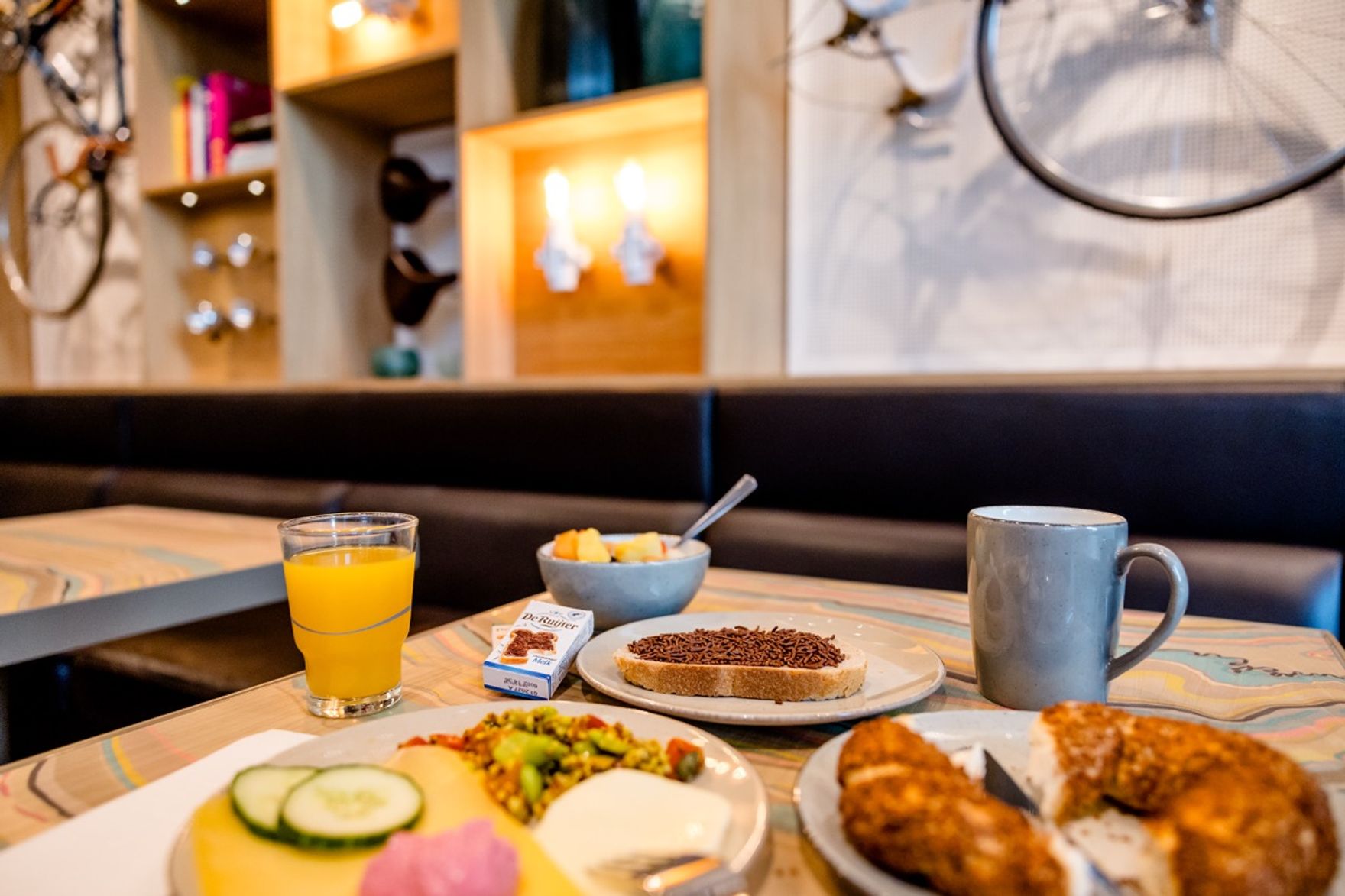 Frühstückstisch mit frisch gepresstem Orangensaft, Müsli, Kaffee, Brot mit Schokostreuseln, Aufschnitt und Gebäck in gemütlichem Café-Ambiente.