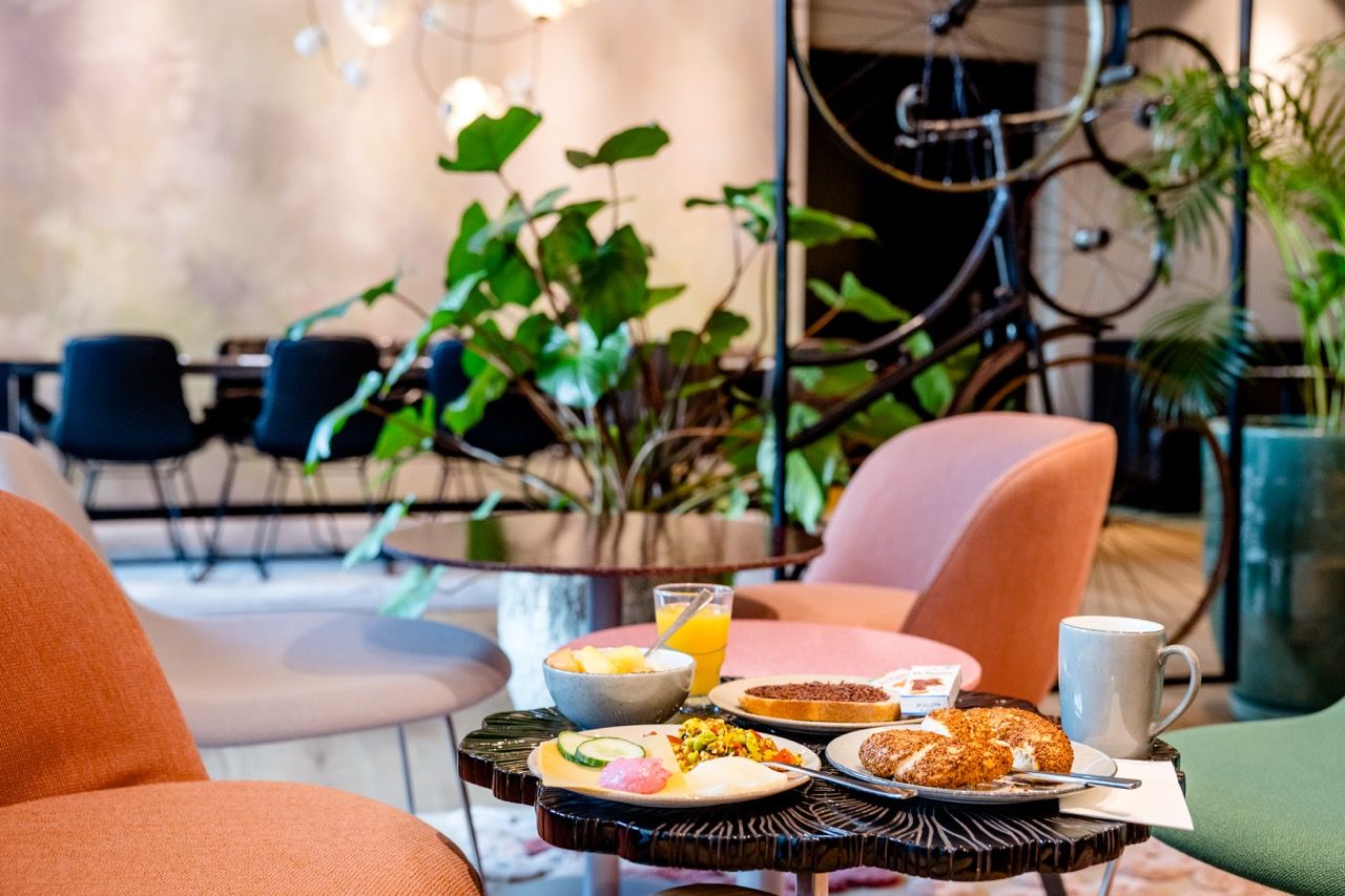 Frühstückstisch mit frischem Gebäck, Obst, Saft und Kaffee in gemütlicher Lounge-Atmosphäre mit bequemen Sesseln und grünen Pflanzen.