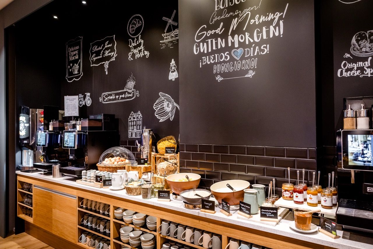 Buffetbereich mit vielfältigem Frühstücksangebot, darunter Brötchen, Marmeladen, Müsli und Kaffeemaschinen, vor schwarzer Wand mit freundlichen morgendlichen Grüßen in verschiedenen Sprachen.