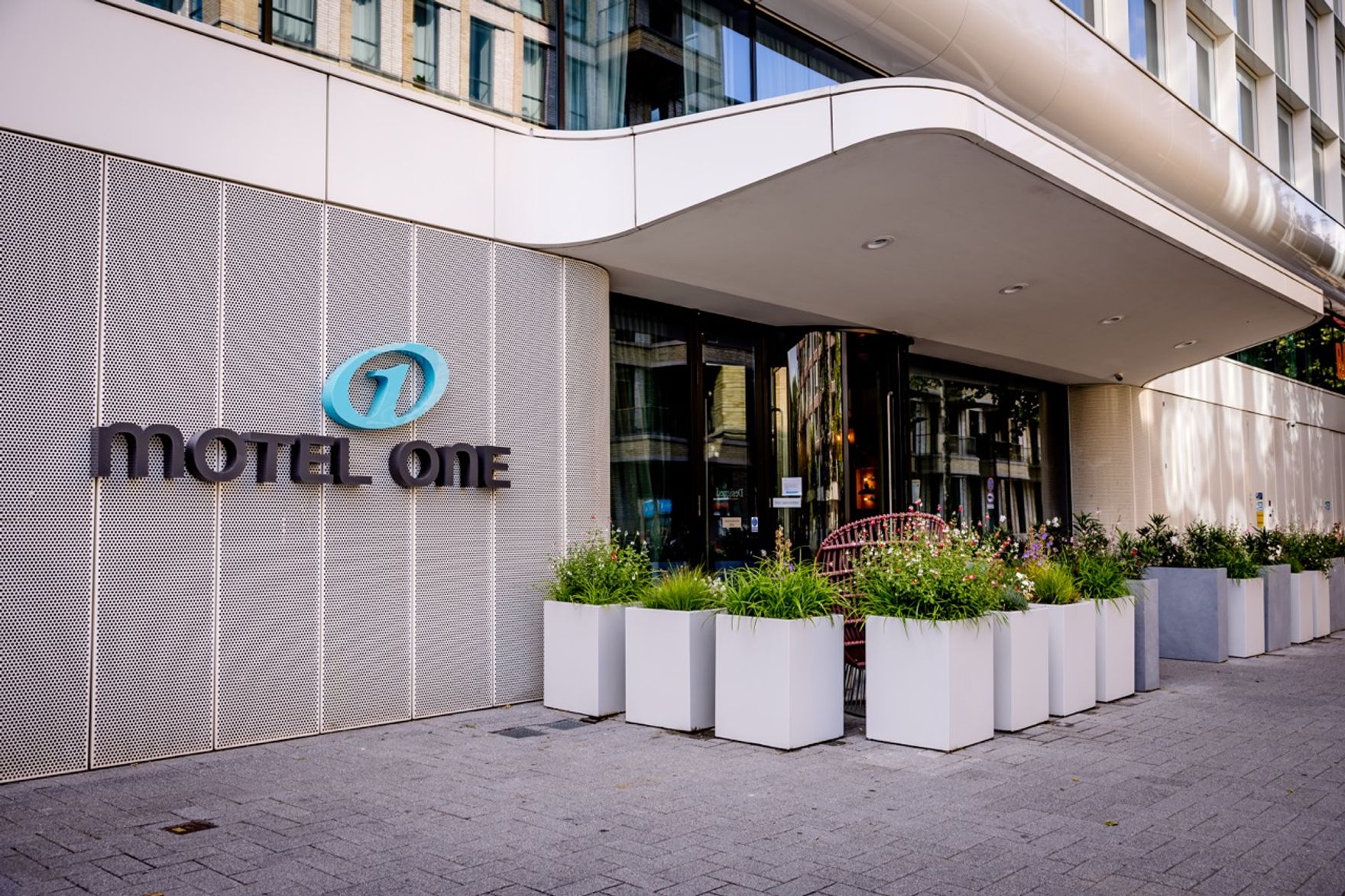 Eingang des Motel One mit modernem Design, großen Glasflächen und Pflanzgefäßen, die den Eingangsbereich freundlich und einladend gestalten.