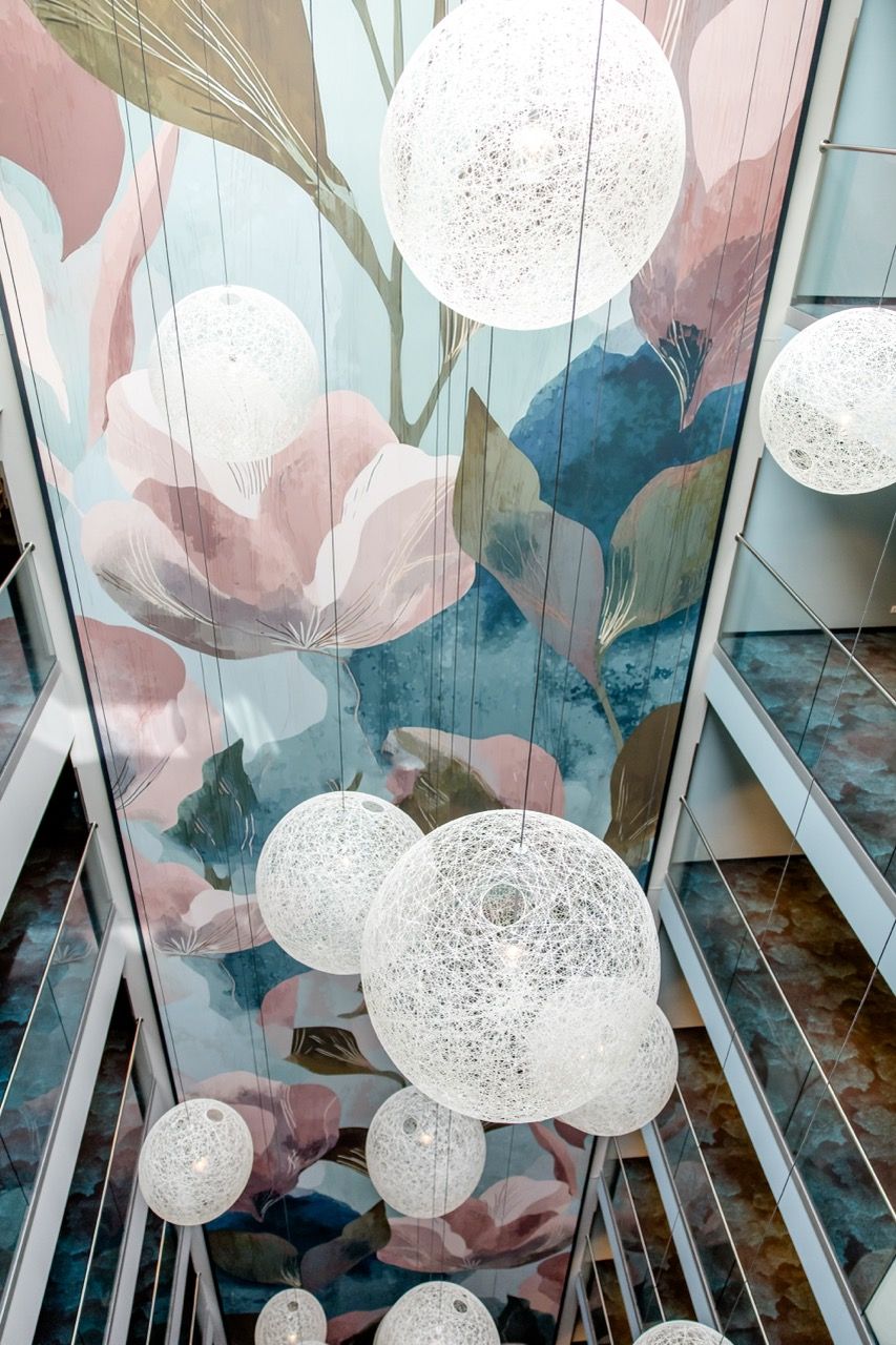Blick nach oben in einen Hotel-Lift-Schacht mit großen, runden, filigranen Hängelampen vor einer farbenfrohen, floralen Wandgestaltung.