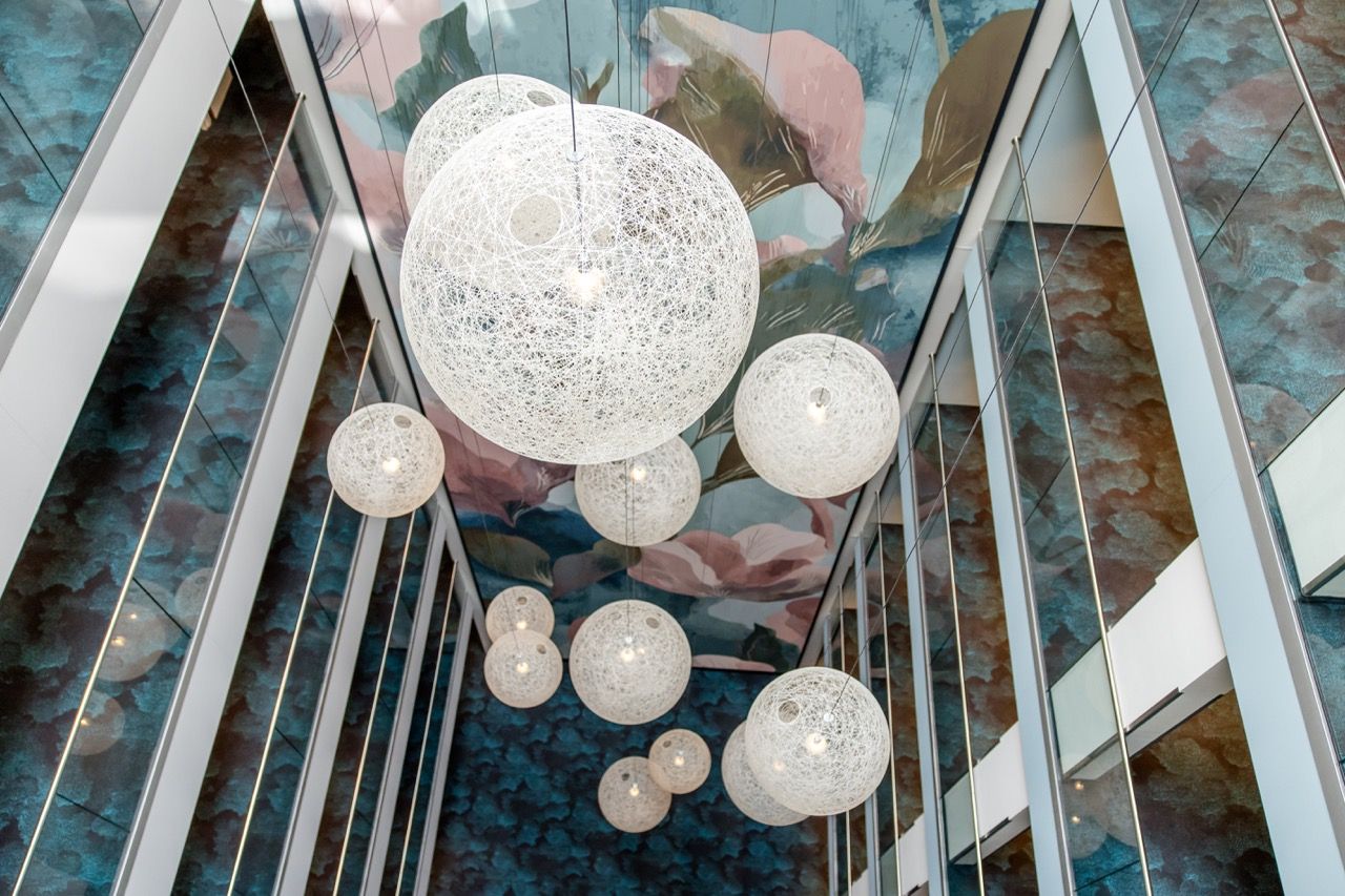 Moderne, kugelförmige Hängeleuchten aus weißem, filigranem Material hängen in unterschiedlicher Größe in einem hohen, lichtdurchfluteten Atrium mit Glaswänden und einer künstlerisch gestalteten Decke.