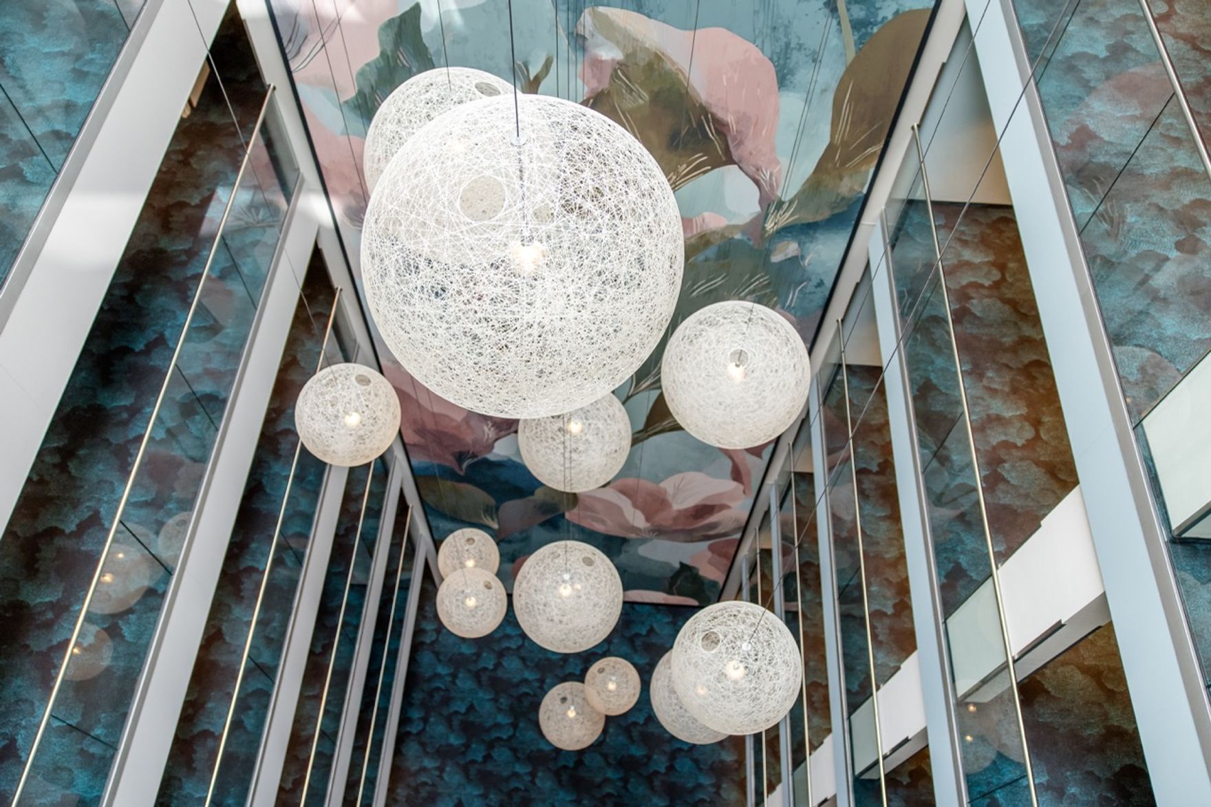 Moderne, kugelförmige Hängeleuchten aus weißem, filigranem Material hängen in unterschiedlicher Größe in einem hohen, lichtdurchfluteten Atrium mit Glaswänden und einer künstlerisch gestalteten Decke.