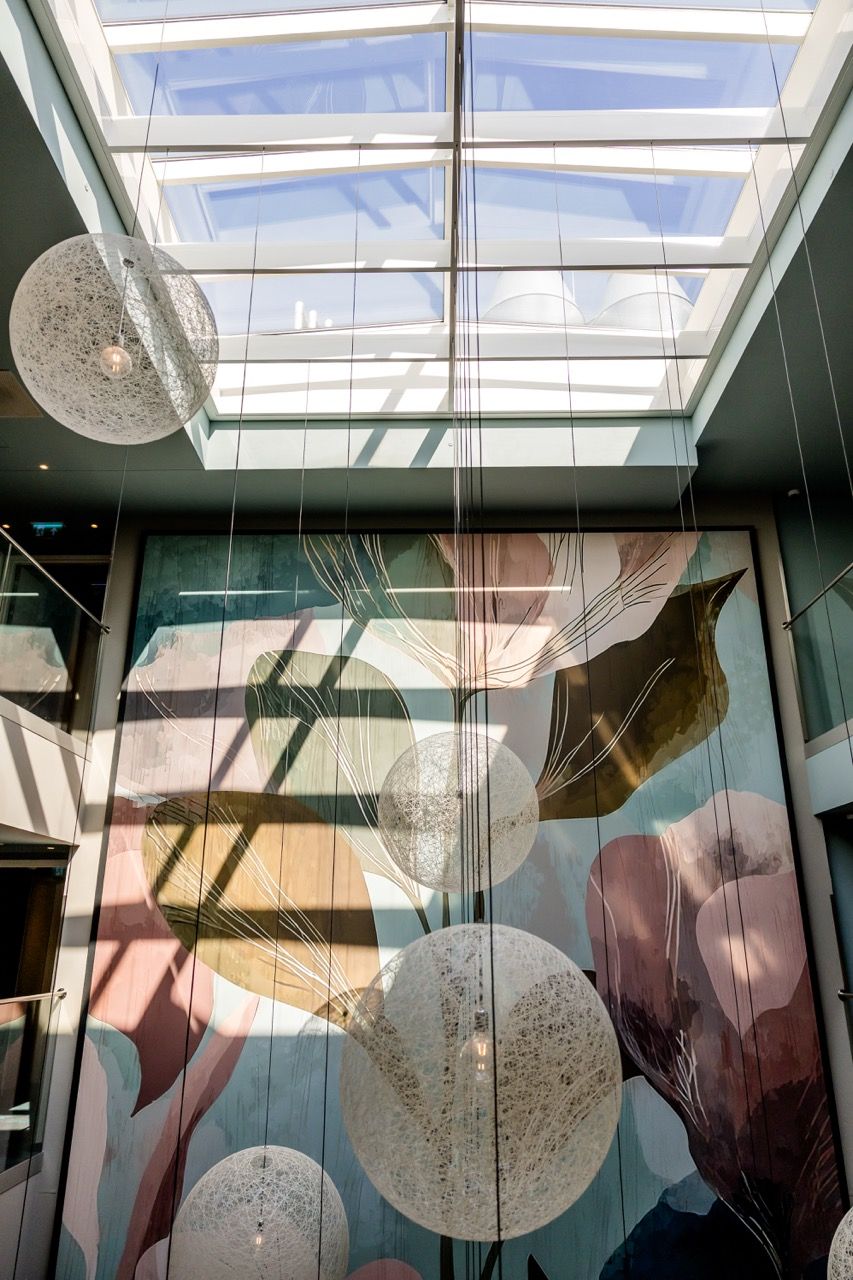 Blick in einen hellen Innenraum mit großer Glasdecke, farbenfrohem Wandbild und modernen, kugelförmigen Hängeleuchten, die leicht in der Luft schweben.
