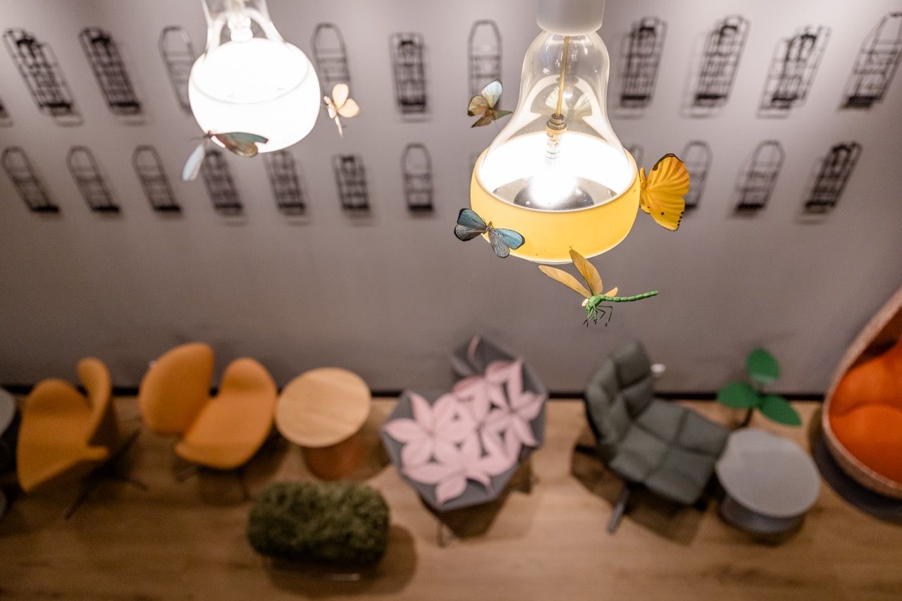 Blick von oben auf eine gemütliche Lounge mit modernen Sesseln, Sofas und kleinen Tischen. Dekorative Schmetterlinge und Libellen umgeben zwei hängende Lampen, die eine warme, einladende Atmosphäre schaffen. Die Wand ist mit stilisierten Fenster-Motiven dekoriert.