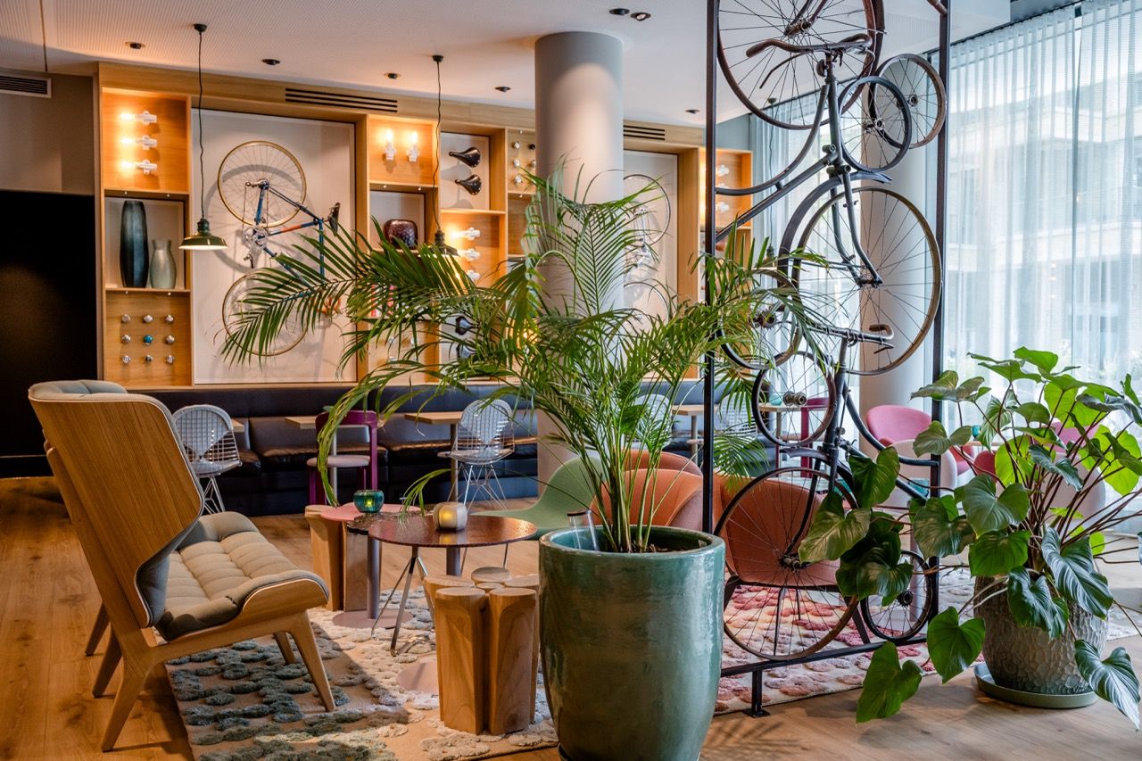 Helle Lounge mit modernen Sitzmöbeln, großen Pflanzen und dekorativ an der Wand und von der Decke hängenden Fahrrädern, die eine entspannte und stilvolle Atmosphäre schaffen.