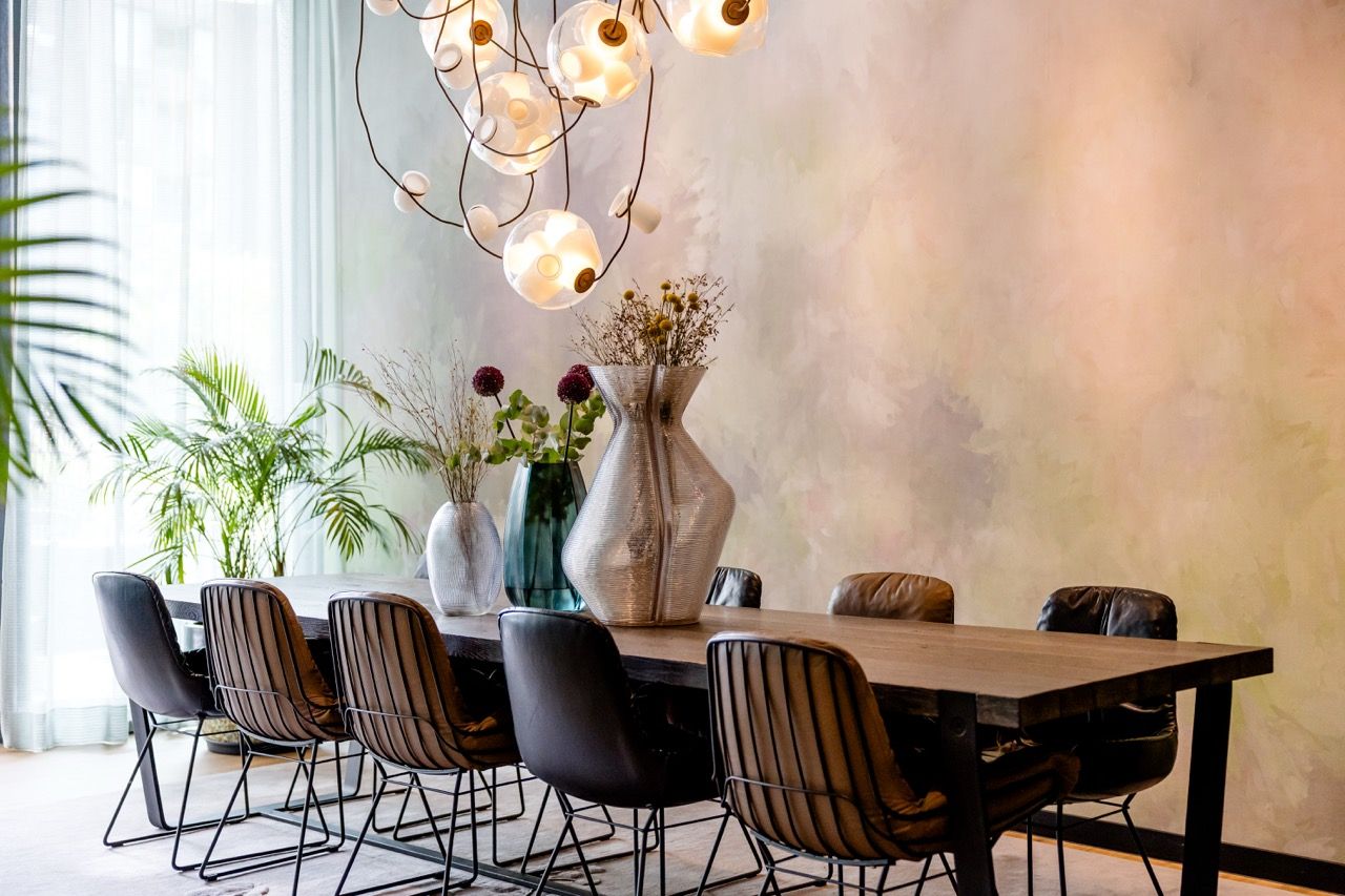 Moderner Essbereich mit langem Holztisch, acht gepolsterten Stühlen und stilvollen Glasvasen mit Blumen. Große Fenster lassen viel Tageslicht herein, warme Pendelleuchten sorgen für gemütliche Atmosphäre. Dezente Wandgestaltung und Pflanzen ergänzen das einladende Raumambiente.