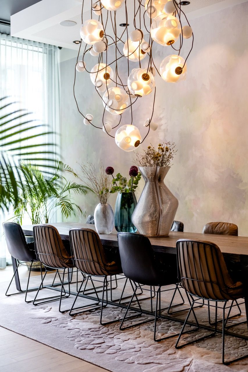 Moderner Essbereich mit langem Holztisch, acht dunklen Stühlen und dekorativen Vasen mit Blumen. Große Fenster lassen Tageslicht herein. Über dem Tisch hängen stilvolle, kugelförmige Pendelleuchten, die warme Atmosphäre schaffen. Pflanzen ergänzen das elegante, einladende Ambiente.