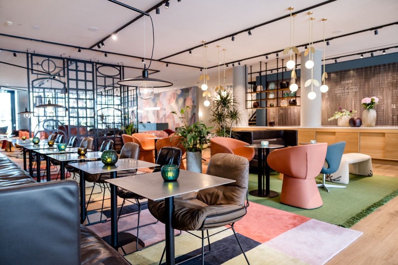Modern eingerichteter Hotel-Lounge-Bereich mit bunten Sesseln, schwarzen Tischen und stilvoller Beleuchtung, der eine entspannte Atmosphäre für Gäste zum Verweilen und Genießen bietet. Pflanzen und großzügige Raumaufteilung schaffen ein einladendes Ambiente.