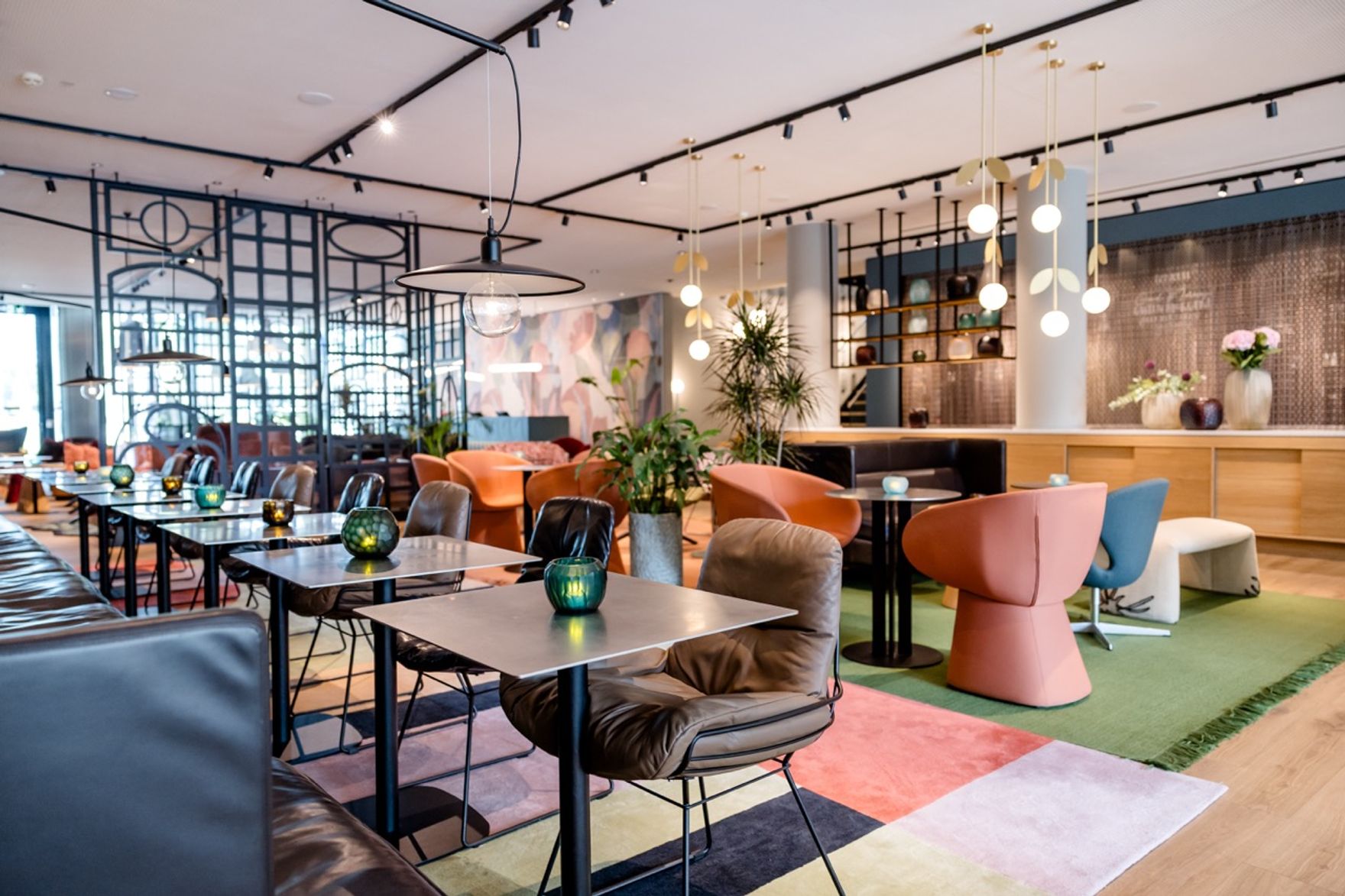 Modern eingerichteter Hotel-Lounge-Bereich mit bunten Sesseln, schwarzen Tischen und stilvoller Beleuchtung, der eine entspannte Atmosphäre für Gäste zum Verweilen und Genießen bietet. Pflanzen und großzügige Raumaufteilung schaffen ein einladendes Ambiente.