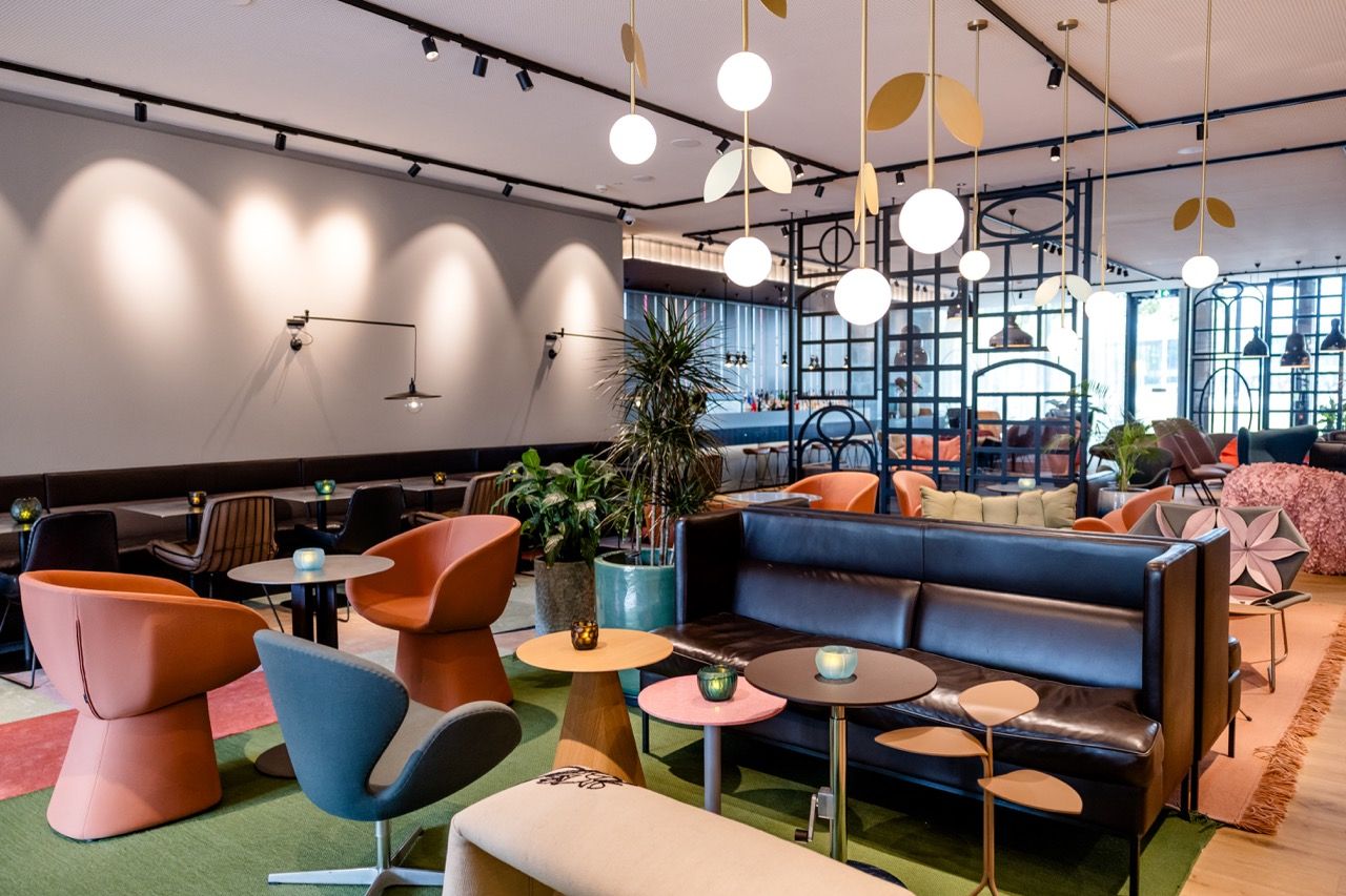 Modernes Hotel-Lounge mit bunten Sesseln, gemütlichen Sofas und stilvollen Tischen. Helle Raumgestaltung, Pflanzen und große Fenster schaffen ein einladendes Ambiente zum Entspannen und Verweilen. Sanfte Beleuchtung mit runden Deckenlampen sorgt für angenehme Atmosphäre.
