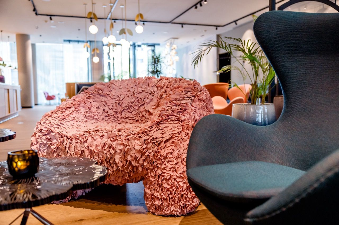 Gemütliche Lounge mit modernem, kreativ gestaltetem rosa Sessel und elegantem dunkelblauem Ohrensessel, umgeben von stilvollen Tischen und Pflanzen, in einem hellen, einladenden Hotelbereich mit großer Fensterfront.