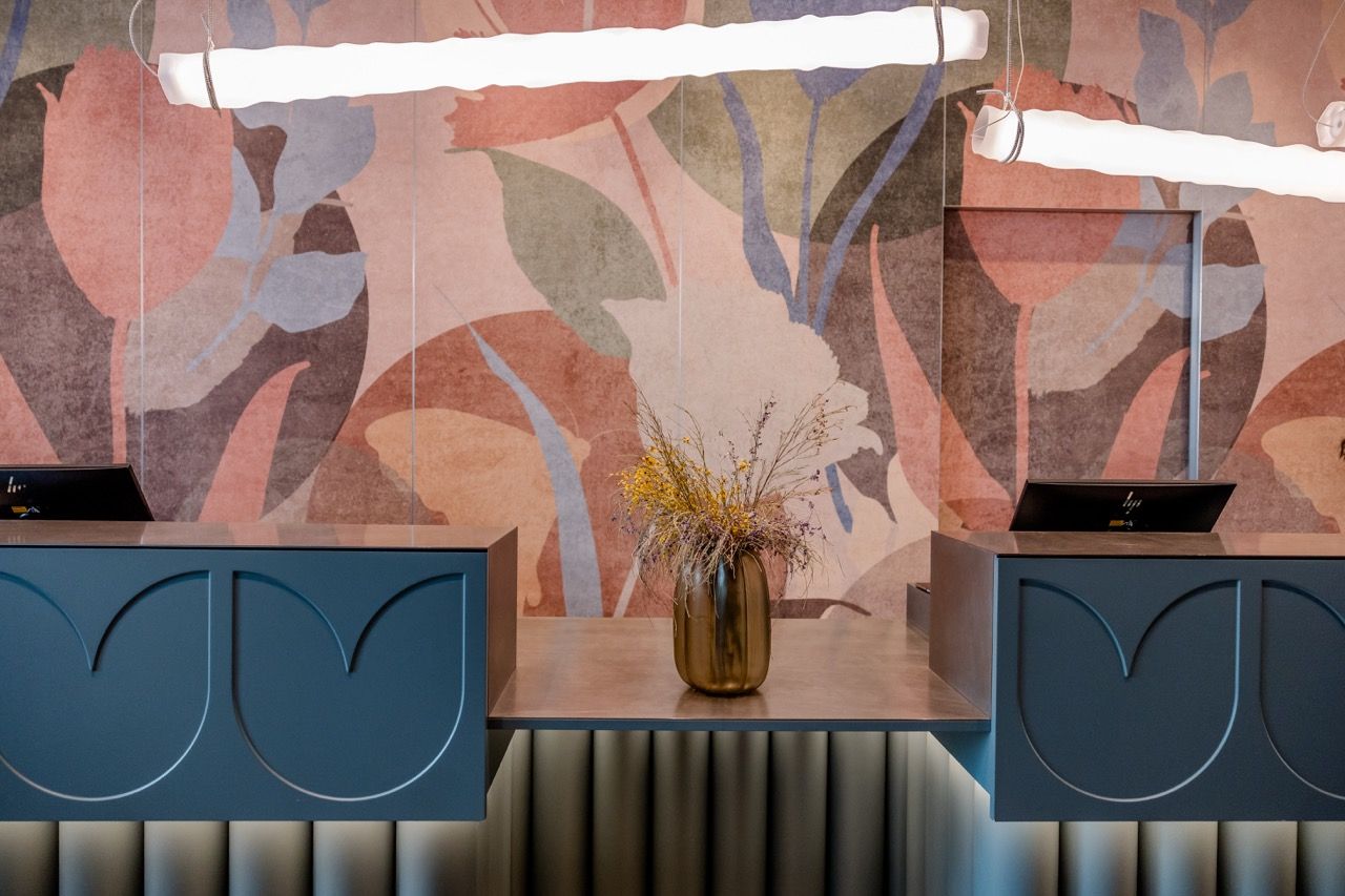 Moderne Hotelrezeption mit dunkelblauen Tresen, stilvoller Beleuchtung und Vase mit Trockenblumen vor dekorativer, floraler Wandgestaltung.