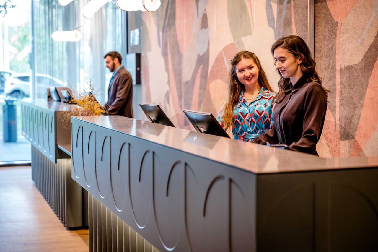 Zwei freundliche Mitarbeiterinnen begrüßen Gäste an einer modernen Hotellobby-Rezeption, im Hintergrund arbeitet ein weiterer Mitarbeiter am Computer.