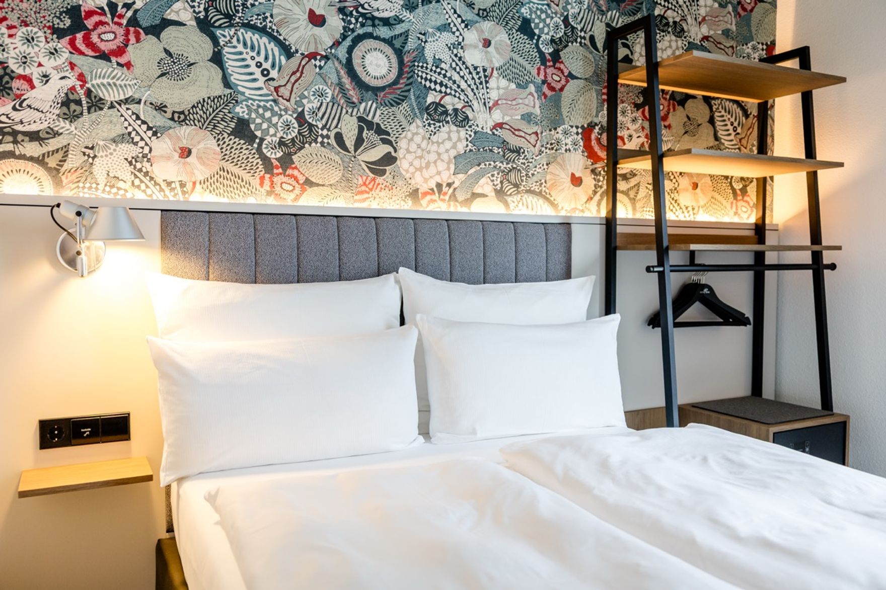 Modern eingerichtetes Hotelzimmer mit gemütlichem Doppelbett, weißen Kissen und Bettwäsche, stilvollem grauem Kopfteil sowie farbenfroher, dekorativer Wandgestaltung im Hintergrund. Neben dem Bett befindet sich eine Wandlampe und ein offenes Regal mit Kleiderbügeln.