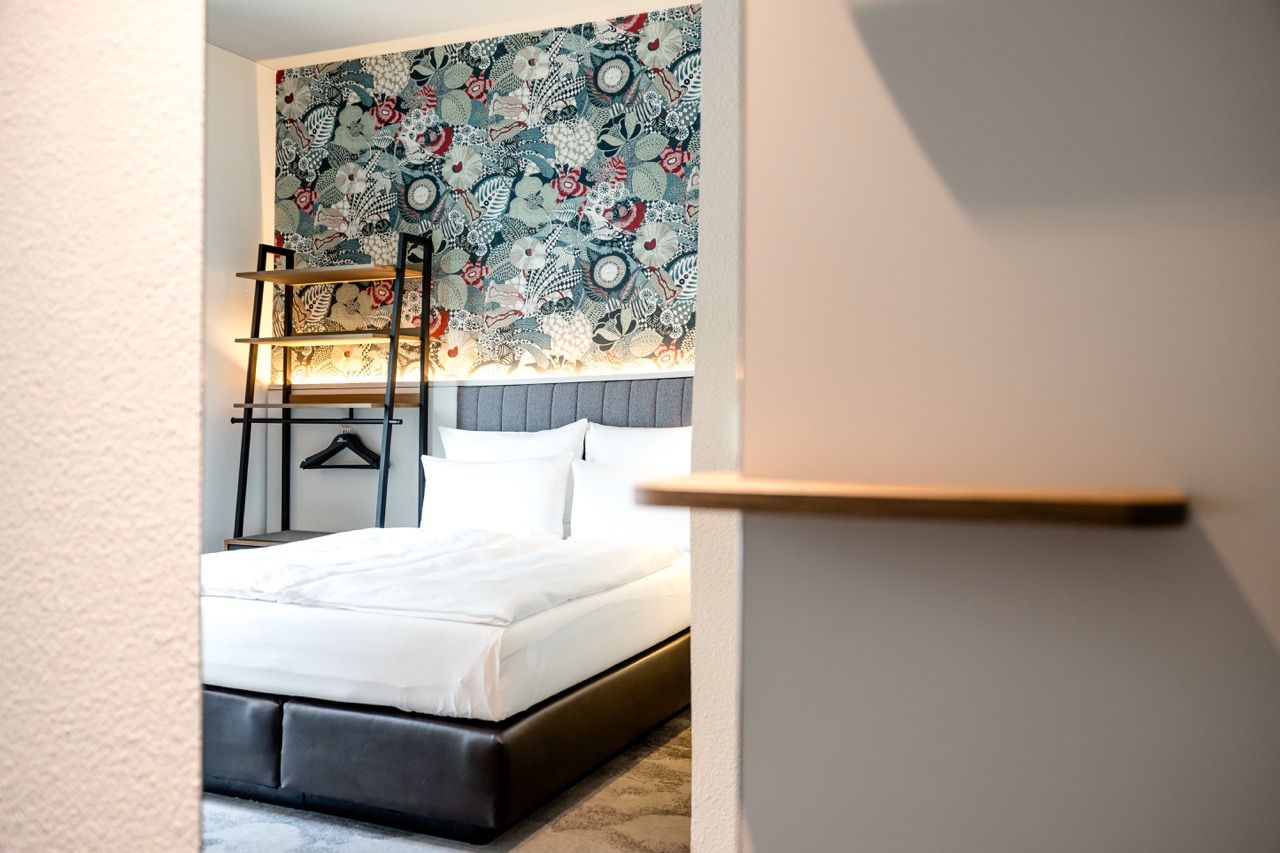 Blick in ein modernes Hotelzimmer mit großem, komfortablem Bett, weißer Bettwäsche und einer dekorativen Blumen- und Blättermuster-Tapete an der Wand, ergänzt durch warme Beleuchtung und einen offenen Kleiderständer.