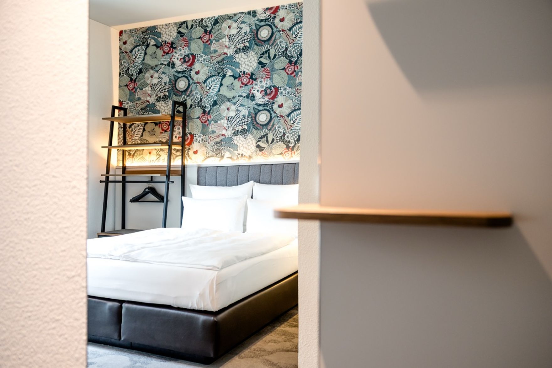 Blick in ein modernes Hotelzimmer mit großem, komfortablem Bett, weißer Bettwäsche und einer dekorativen Blumen- und Blättermuster-Tapete an der Wand, ergänzt durch warme Beleuchtung und einen offenen Kleiderständer.