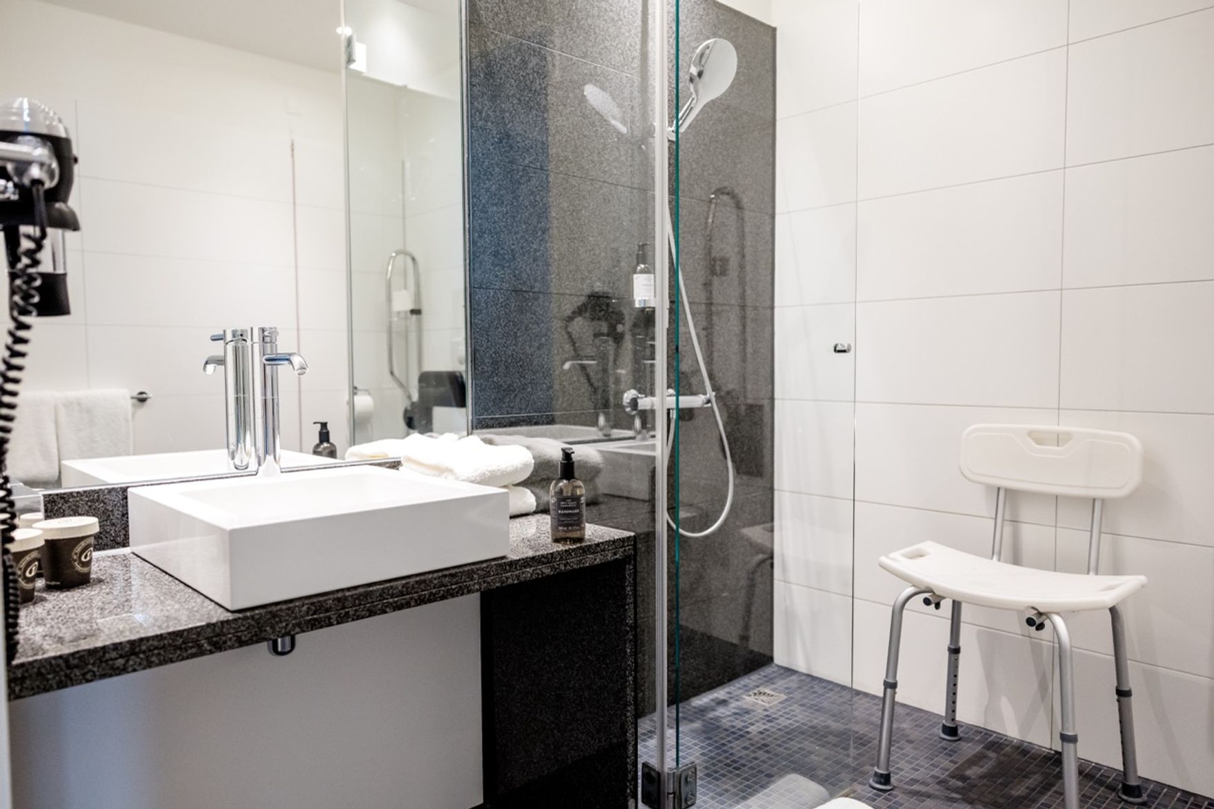 Modernes Badezimmer mit barrierefreier Dusche, Duschstuhl und großem Spiegel. Helle, freundliche Atmosphäre mit klaren Linien und funktionaler Ausstattung.