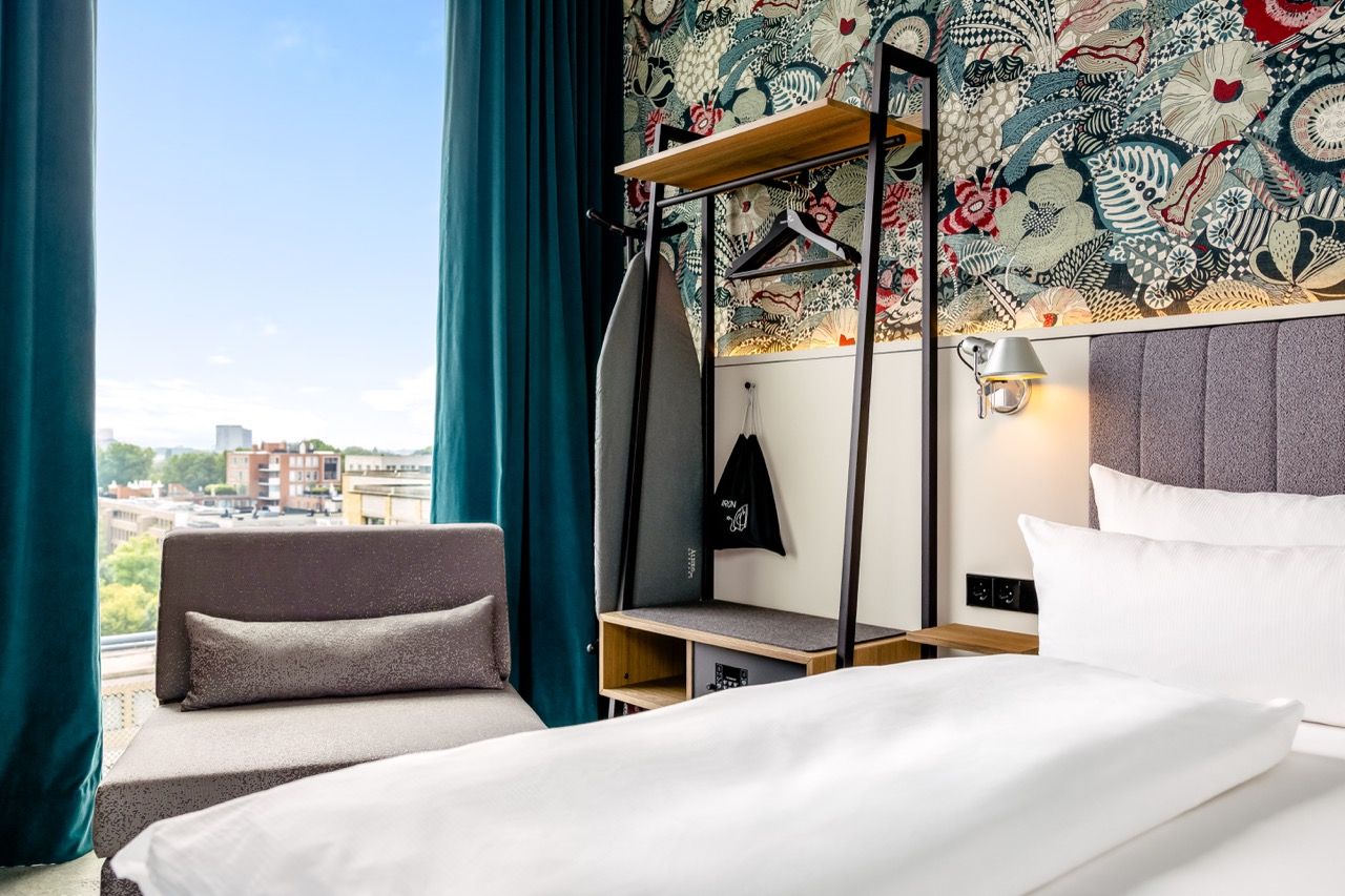 Modern eingerichtetes Hotelzimmer mit gemütlichem Doppelbett, Sitzgelegenheit am Fenster und beranktem Wandregal, das funktionalen Stauraum bietet. Große Fenster mit Blick auf die Stadt und florale Tapete schaffen eine helle, einladende Atmosphäre.