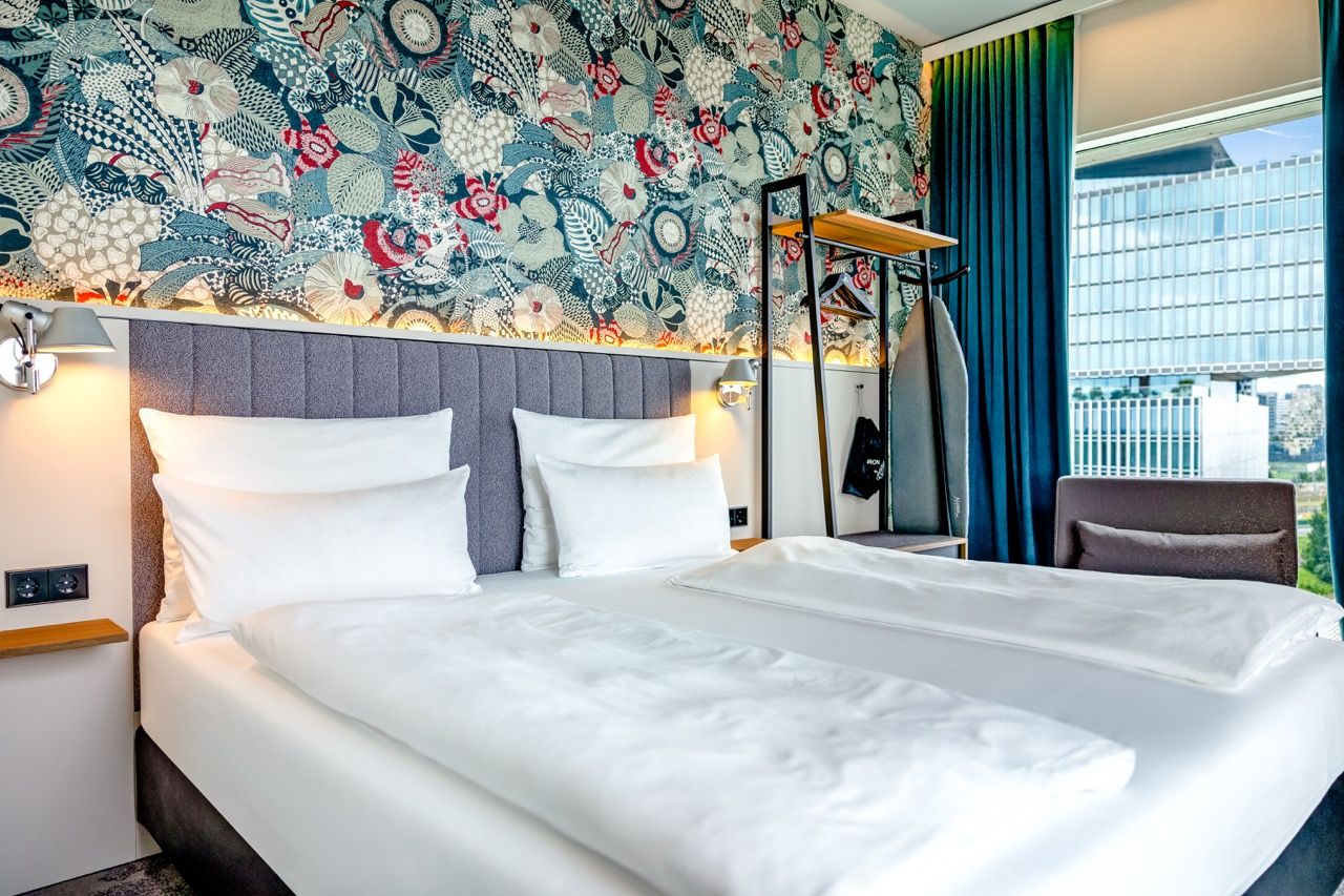 Modernes Hotelzimmer mit zwei frisch bezogenen Einzelbetten, dekorativer Wandtapete und großer Fensterfront mit Blick auf Stadtgebäude, das Tageslicht hereinlässt. Ein eleganter, gemütlicher Ort für erholsamen Aufenthalt und entspannte Nächte.