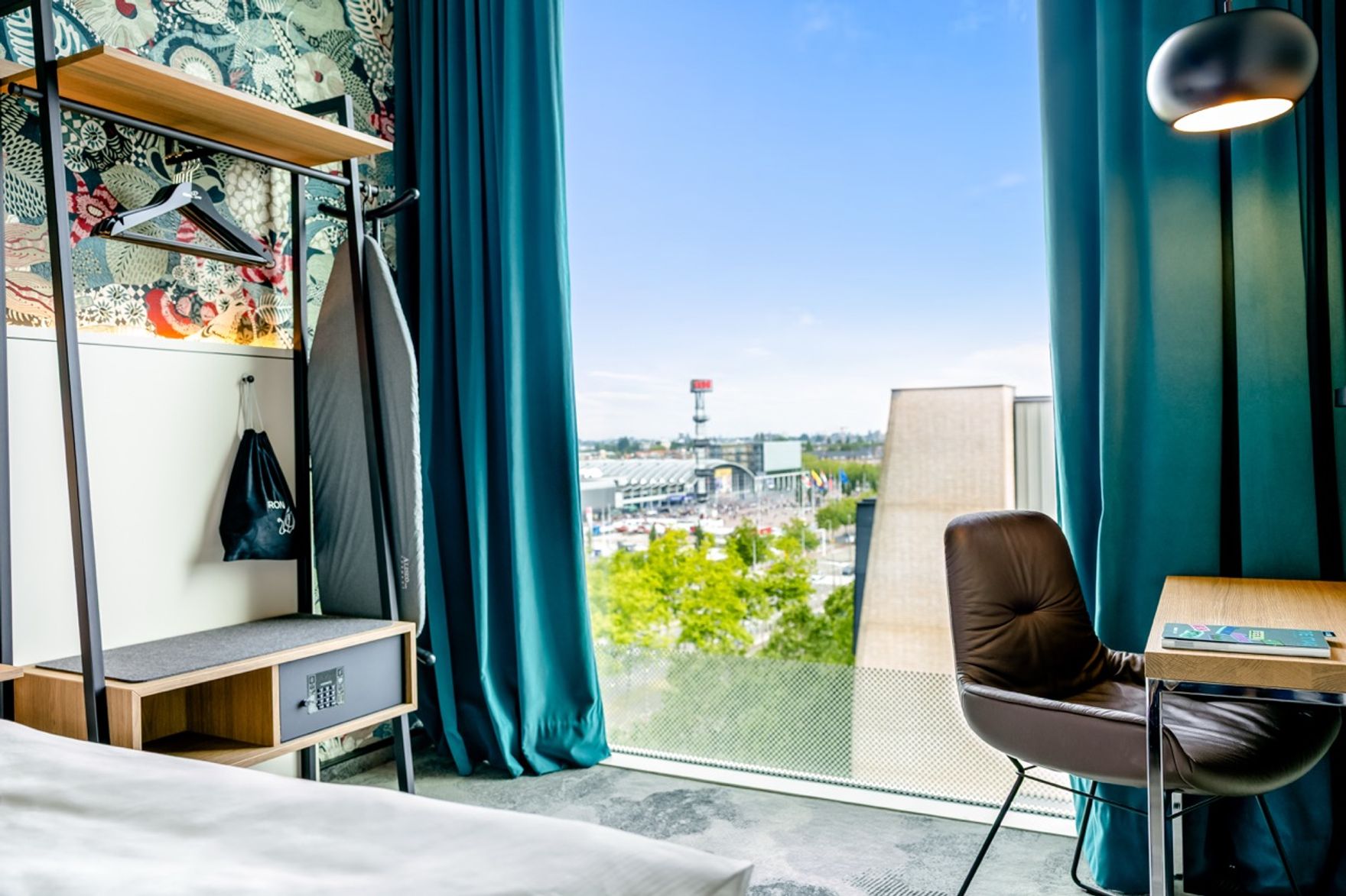 Modern eingerichtetes Hotelzimmer mit großem Fenster, das eine weite Aussicht auf die Stadt und grüne Bäume bietet. Helles Tageslicht, Sitzbereich mit Stuhl und Schreibtisch, gemütliche Atmosphäre durch farbige Vorhänge und stilvolle Dekoration.