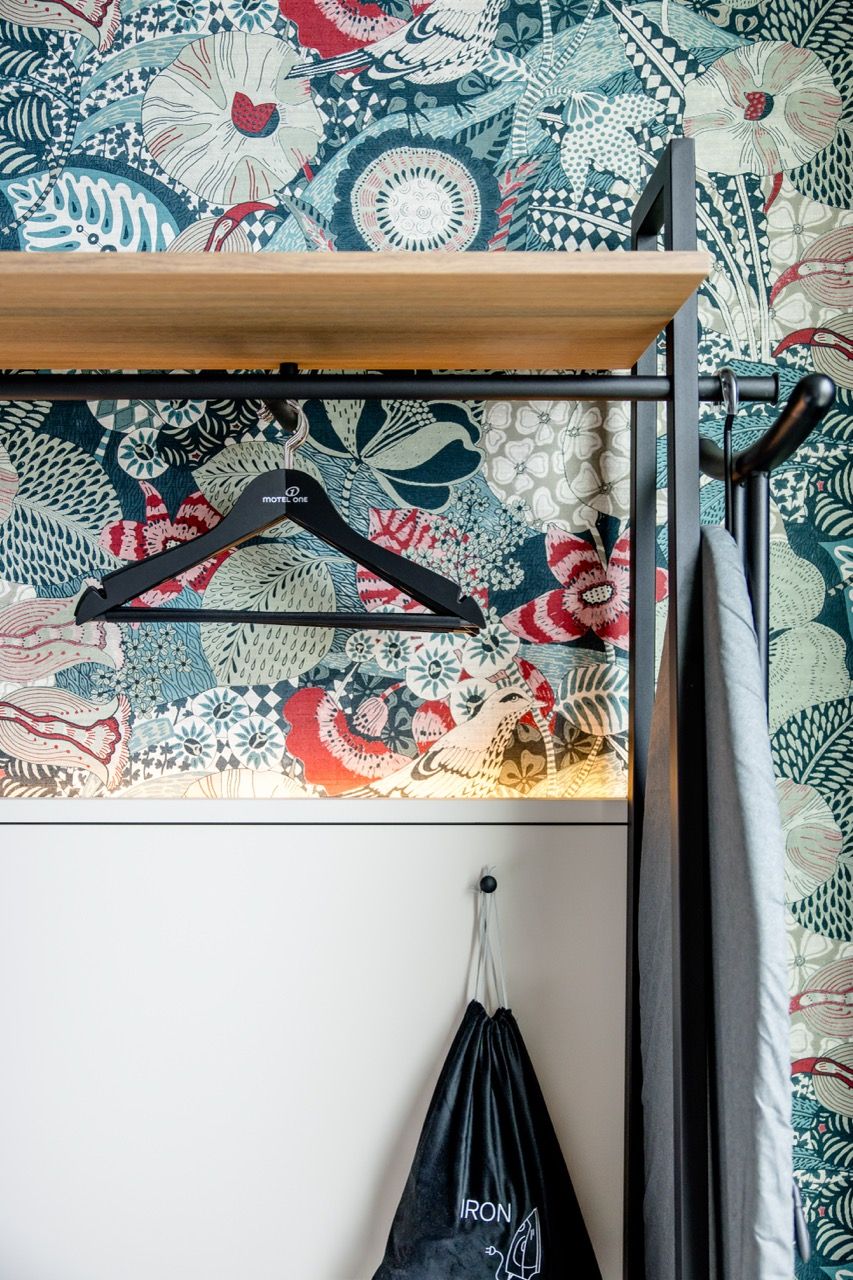 Offene Garderobe mit buntem Blumenmuster an der Wand, einer Holzablage, schwarzen Kleiderbügeln und hängendem Wäschebeutel. Modern und einladend gestaltet.