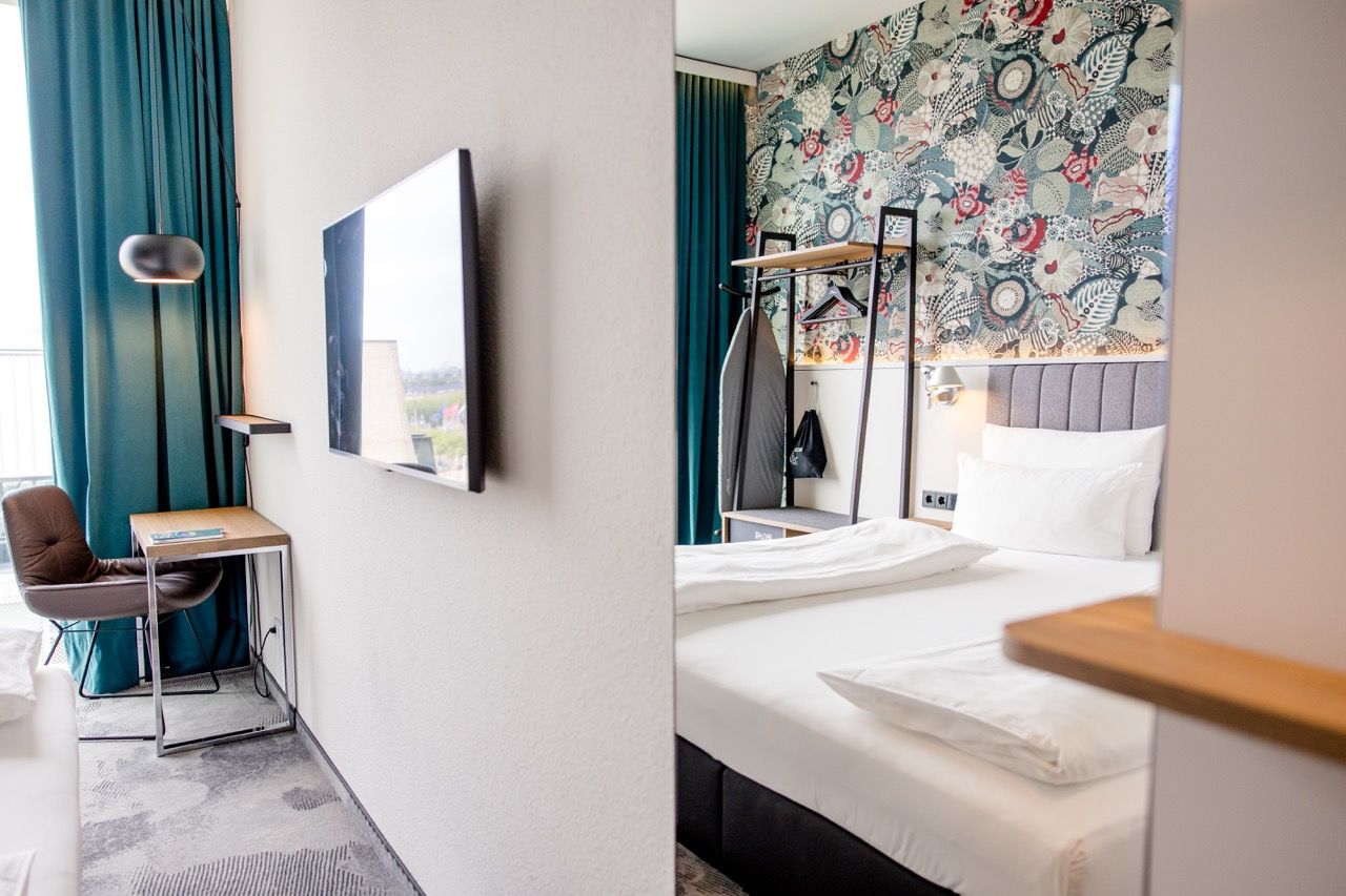 Helles, modernes Hotelzimmer mit großem Doppelbett, bunter Musterwand, Schreibtisch, Flachbildfernseher und dunklen Vorhängen, das eine gemütliche und stilvolle Atmosphäre bietet.