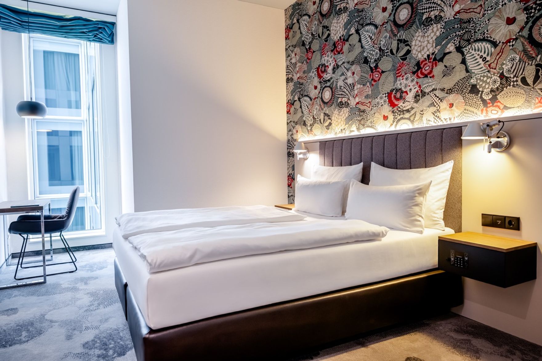 Modernes Hotelzimmer mit großem, komfortablem Doppelbett, dekorativer Wandgestaltung, Schreibtisch am Fenster und sanfter Beleuchtung für eine entspannte Atmosphäre.