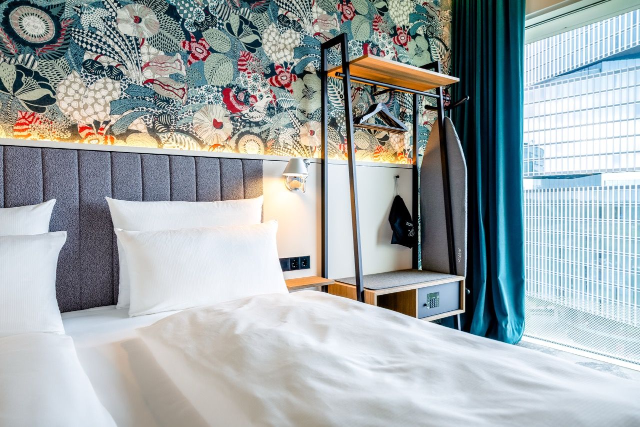 Modern eingerichtetes Hotelzimmer mit gemütlichem Doppelbett, farbenfroher Wandgestaltung, offenem Kleiderständer und großem Fenster mit blickdichten Vorhängen, das helles Tageslicht in den Raum lässt.