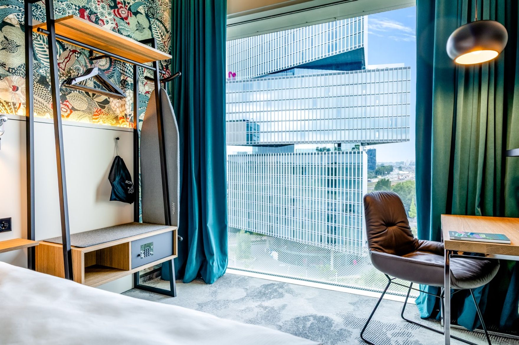 Hotelzimmer mit großem Fenster und Blick auf moderne Hochhäuser, gemütlichem Sessel am Schreibtisch, Garderobe mit Haken, Safe und dekorativer Wand. Helle, freundliche Atmosphäre mit türkisfarbenen Vorhängen.