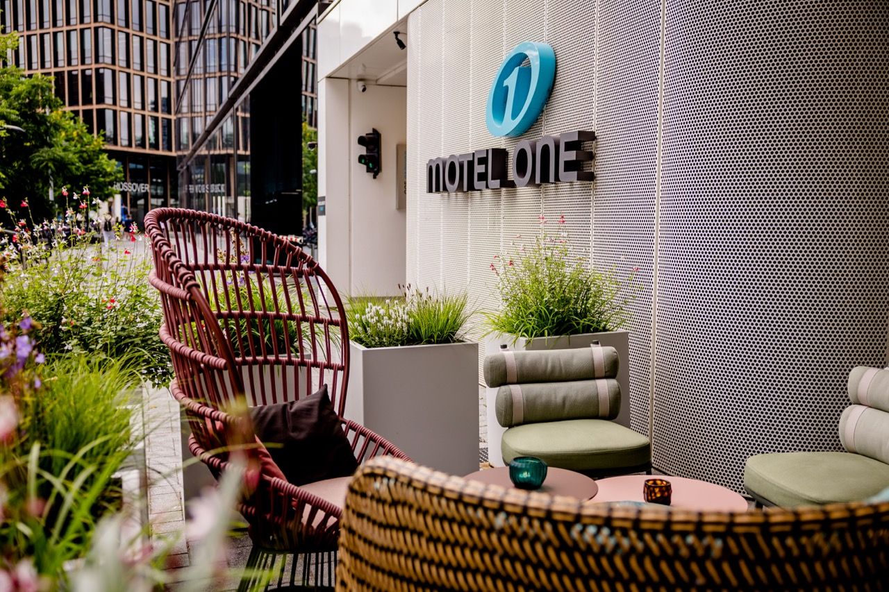 Außenbereich mit gemütlichen Sitzmöbeln und Pflanzen vor dem modernen Motel One Gebäude, ideal für entspannte Pausen im urbanen Umfeld.